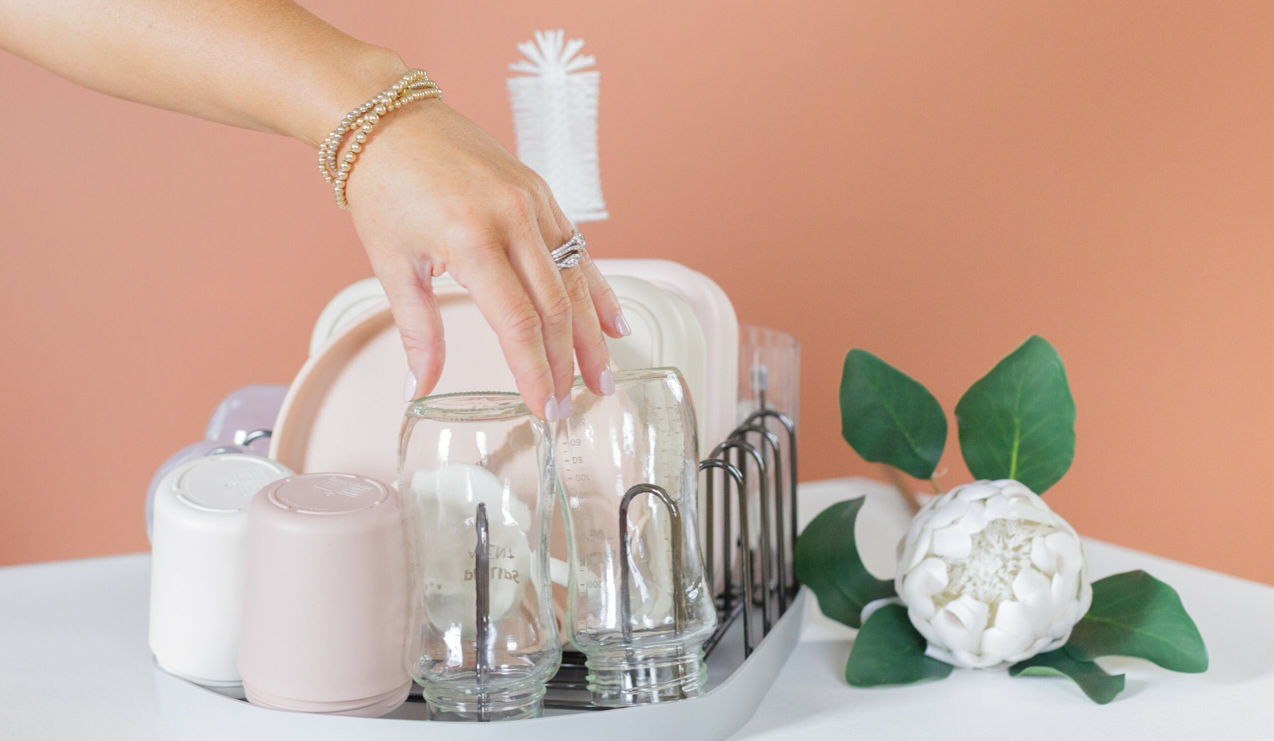 Hand placing a glass on a tray with other glasses, white flower with green leaves nearby, peach background.