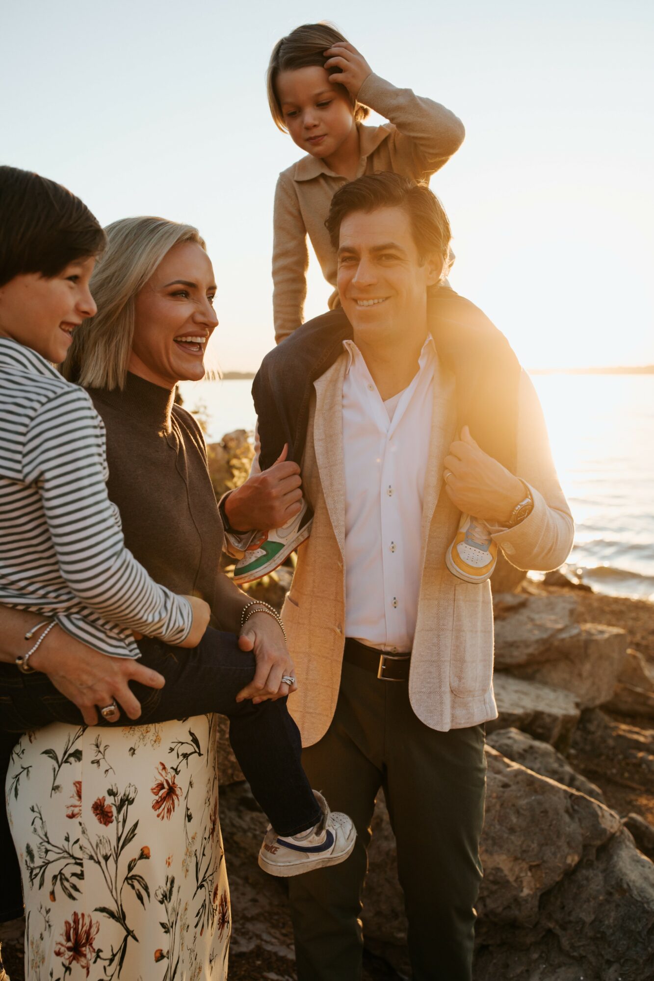 Group of five people outdoors near water, smiling, with a sunset background, one person sitting on a rock, others standing.