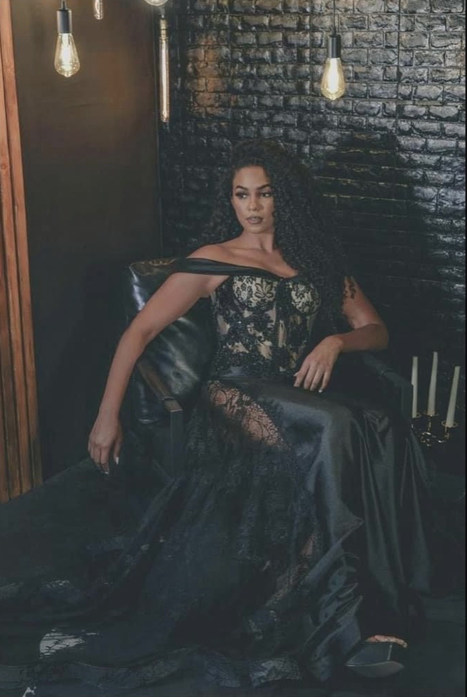 Woman in black lace dress sitting on a black leather chair against a dark brick wall with hanging light bulbs.