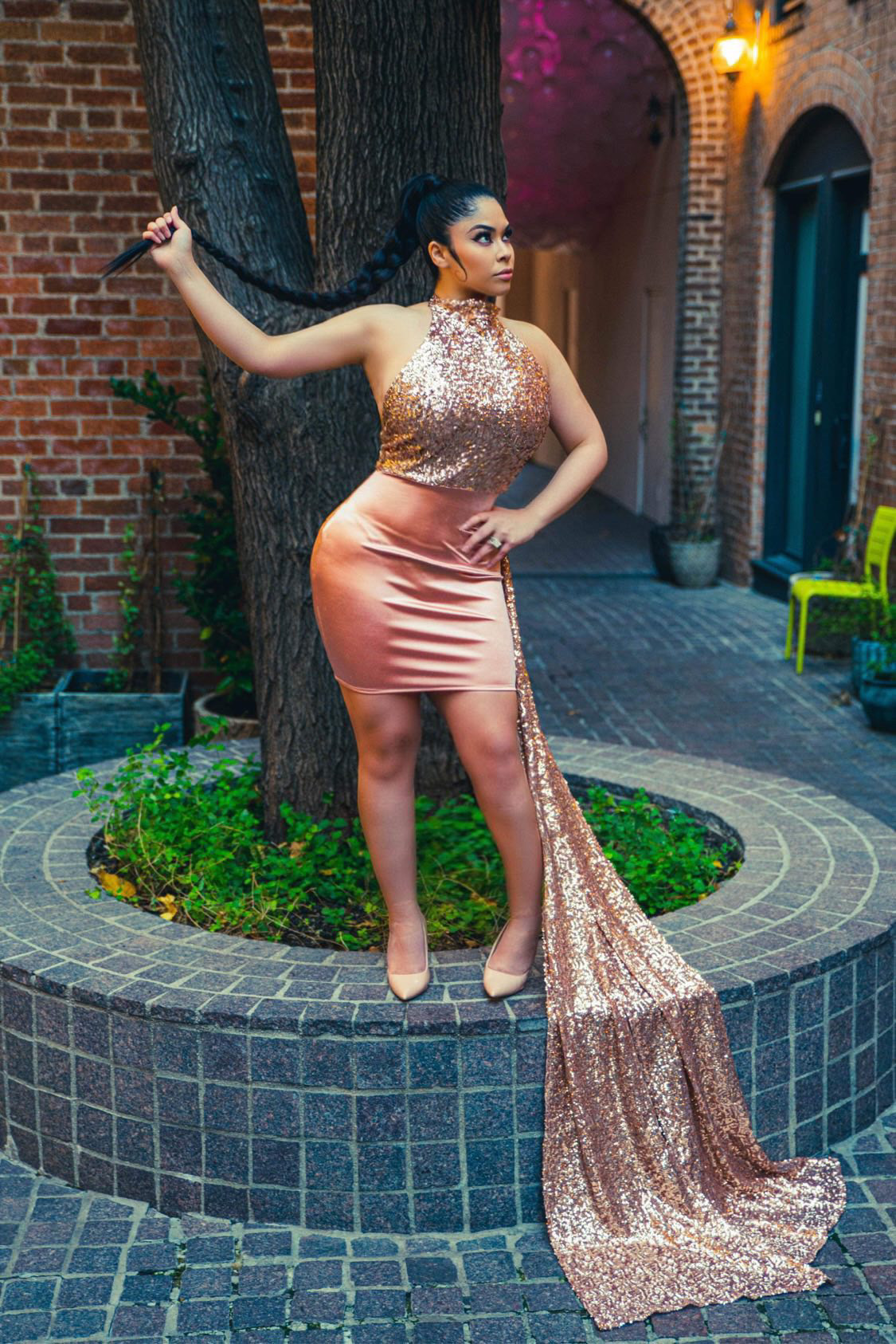 Woman in a shiny, two-tone dress with a long, flowing train, standing outdoors near a tree and brick building.