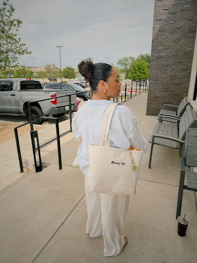 Woman with dark hair in a bun, wearing light-colored clothing, carrying a tote bag, standing outside near chairs and a building.