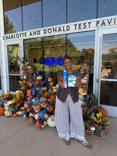 Smiling woman holding a certificate and a drink standing in front of a decorated entrance with flowers, glass doors, and a sign.