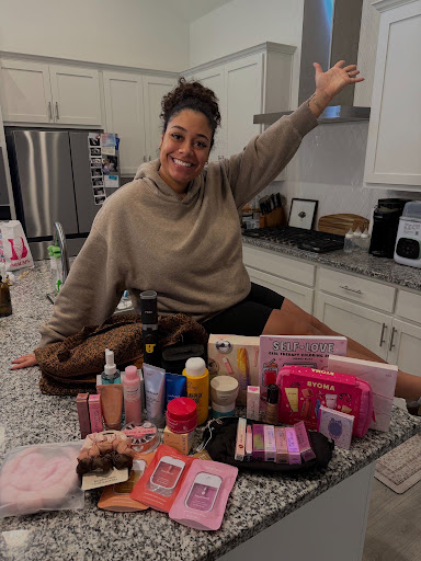 Smiling woman with curly hair in beige sweater sitting on kitchen counter, raising arm, surrounded by skincare products and pink boxes.