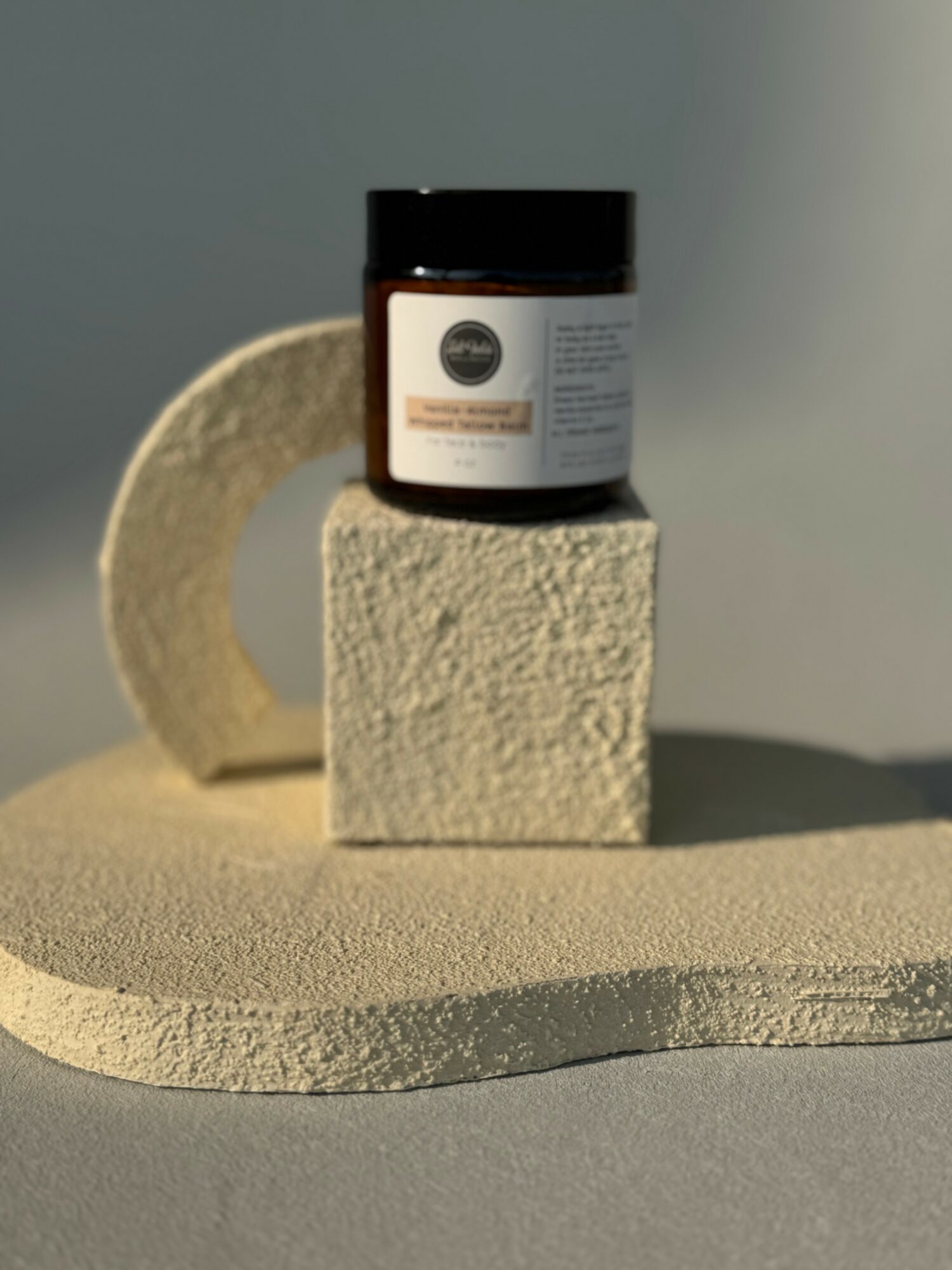 A small jar with a black lid is placed on a textured block, which is on a curved surface with a shadowed background.