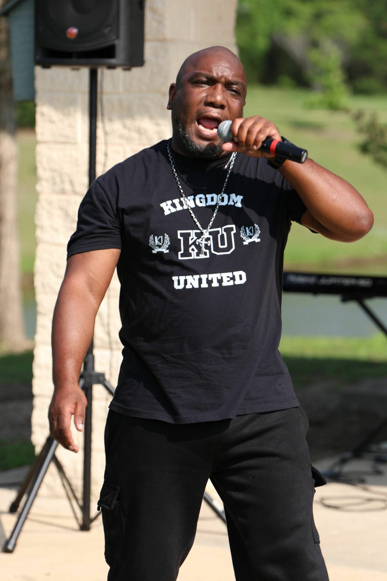 Person singing into a microphone outdoors, wearing a black T-shirt and black pants, with a speaker in the background.