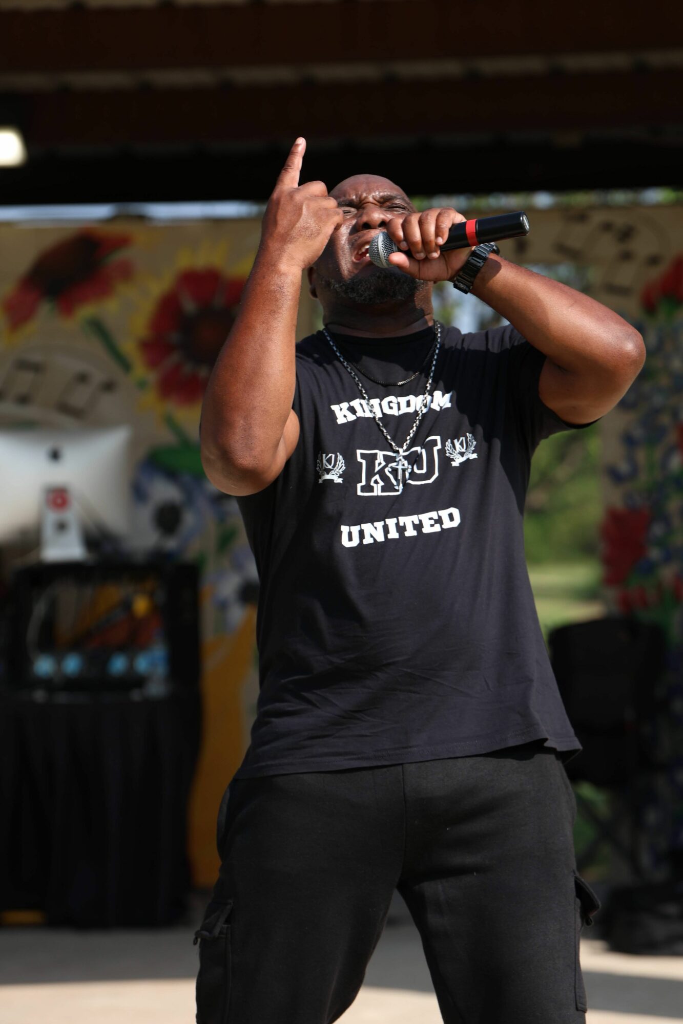 Person holding a microphone and pointing with their right hand, wearing a black T-shirt with white text, in front of a colorful background.