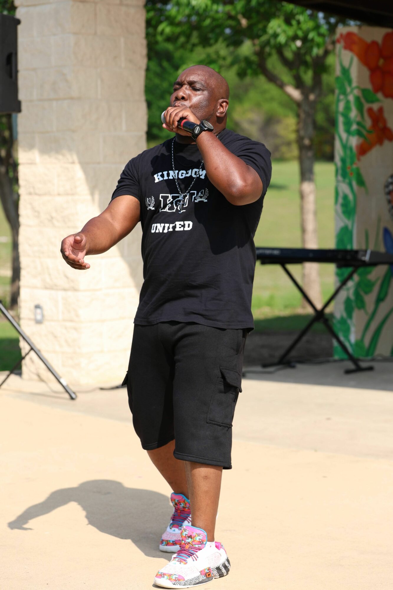 Person speaking into a microphone outdoors, wearing black clothing and sunglasses, with trees and a colorful banner in background.
