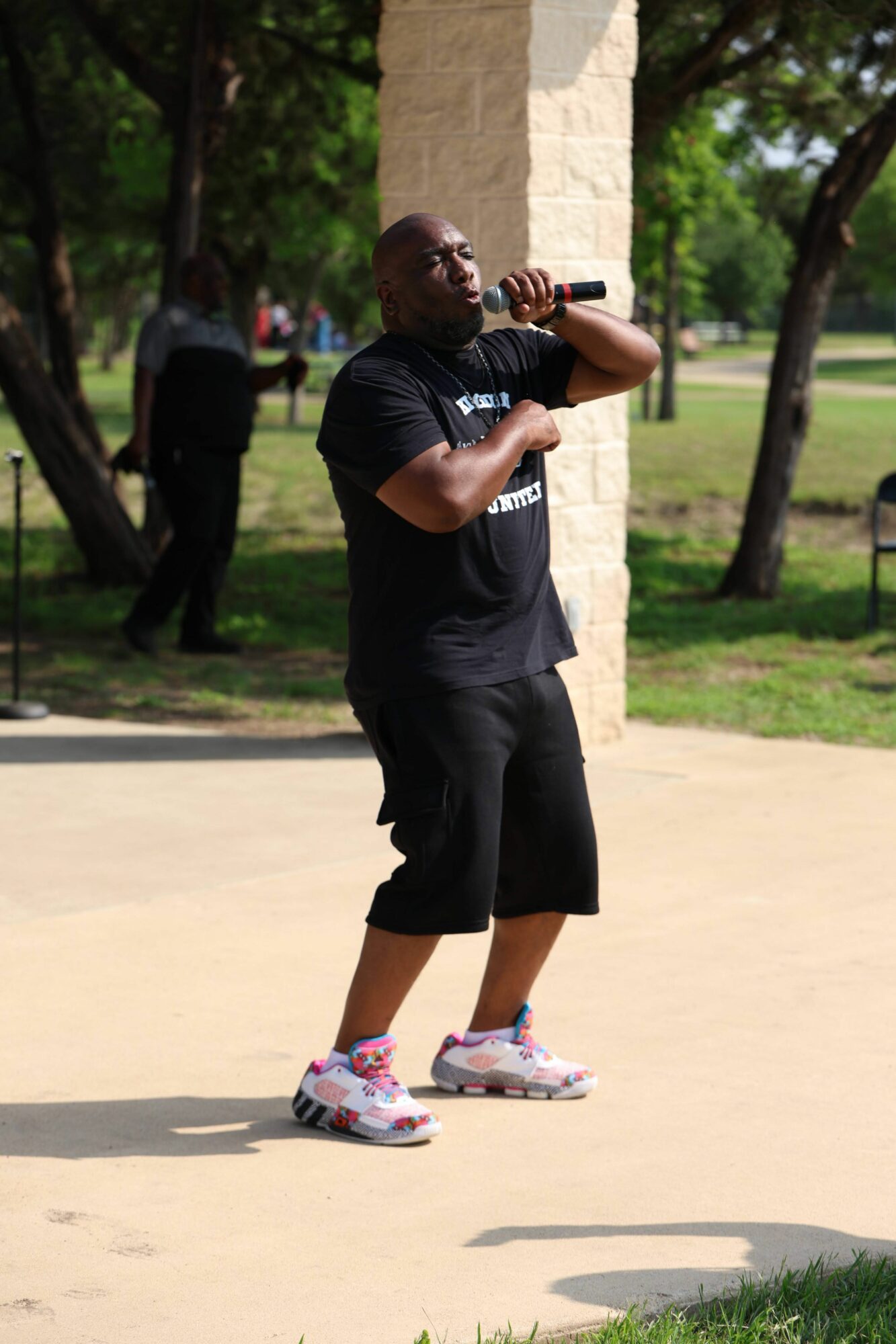 Person speaking into a microphone outdoors, holding a paper, with trees and a stone structure in the background.