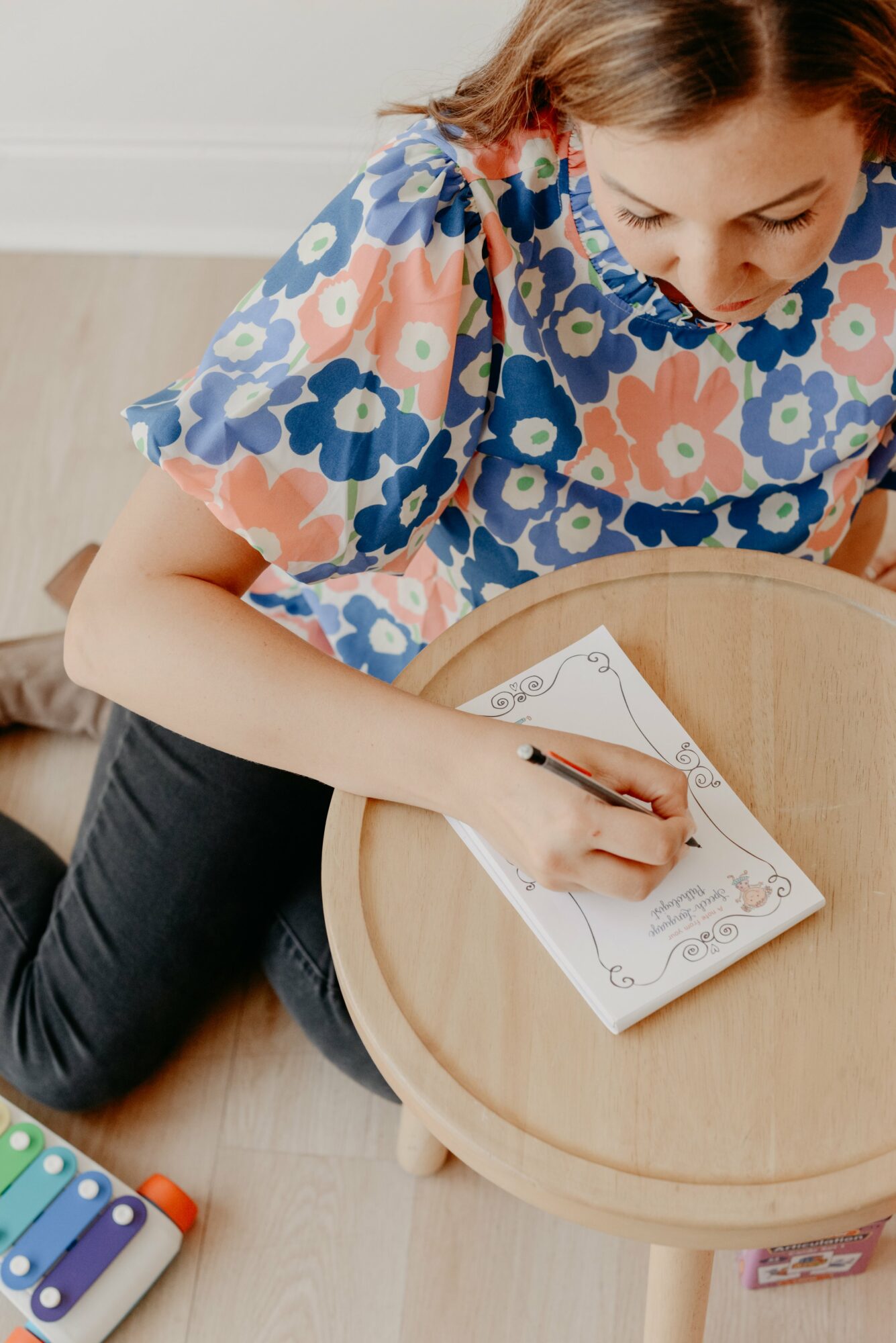 Child writing on a paper with a pen, sitting at a round wooden table, wearing a colorful floral shirt.