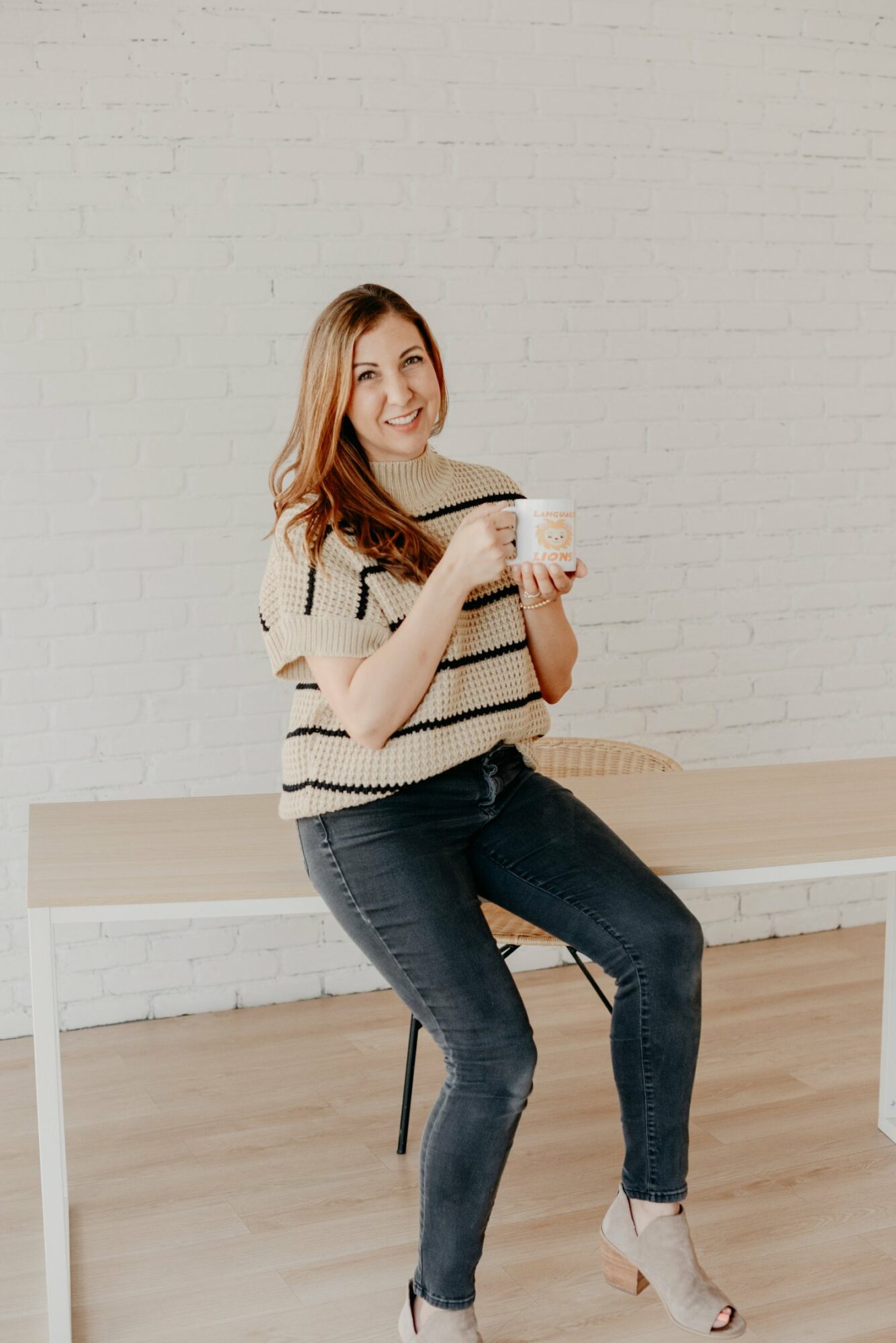 Young woman sitting on a bench, smiling, holding a mug, against a white brick wall, casual clothing, light-colored sweater, jeans, heels.