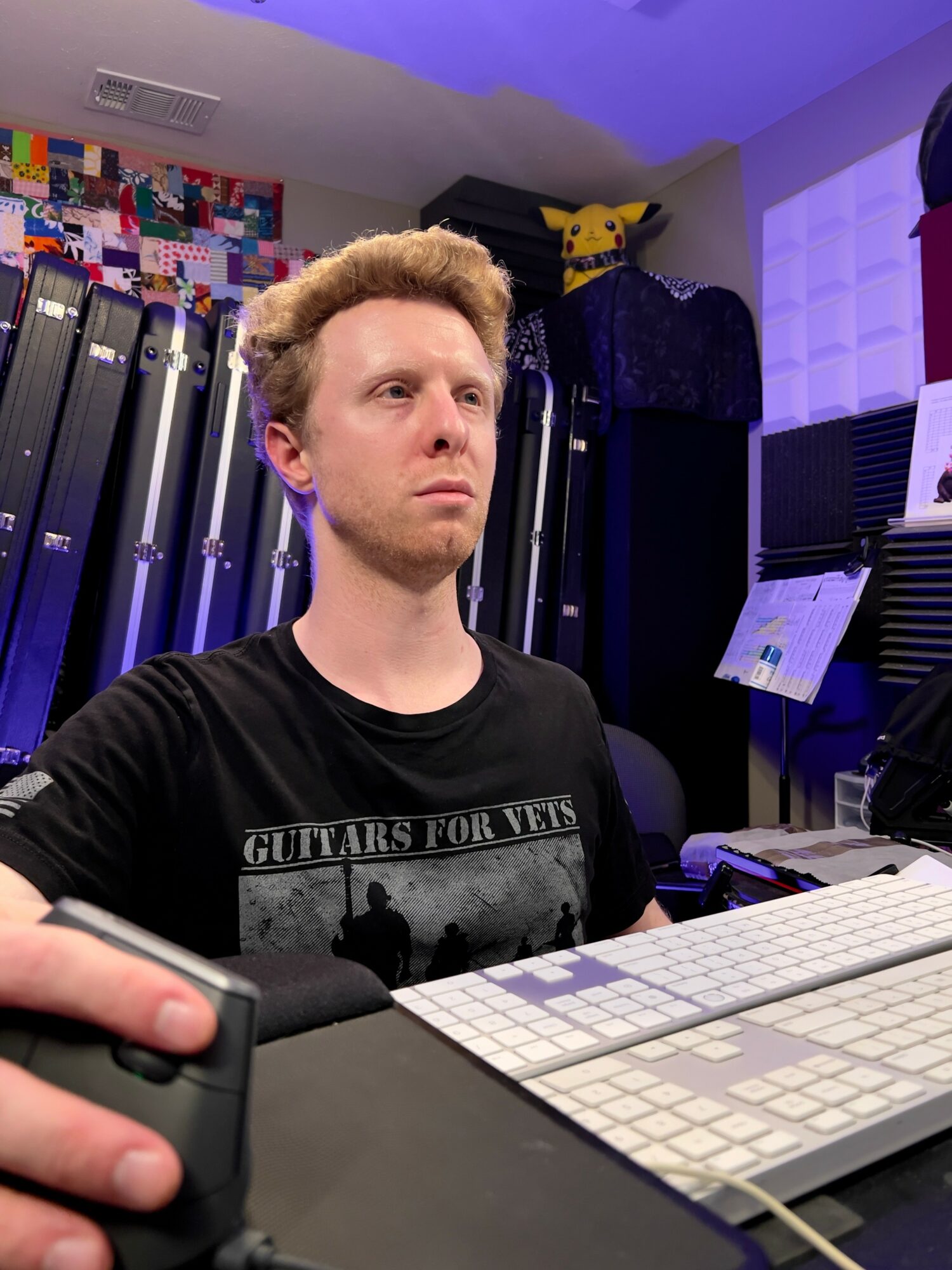 Young man with curly hair sitting at a desk with a keyboard and monitor, in a room with colorful wall decor and equipment.