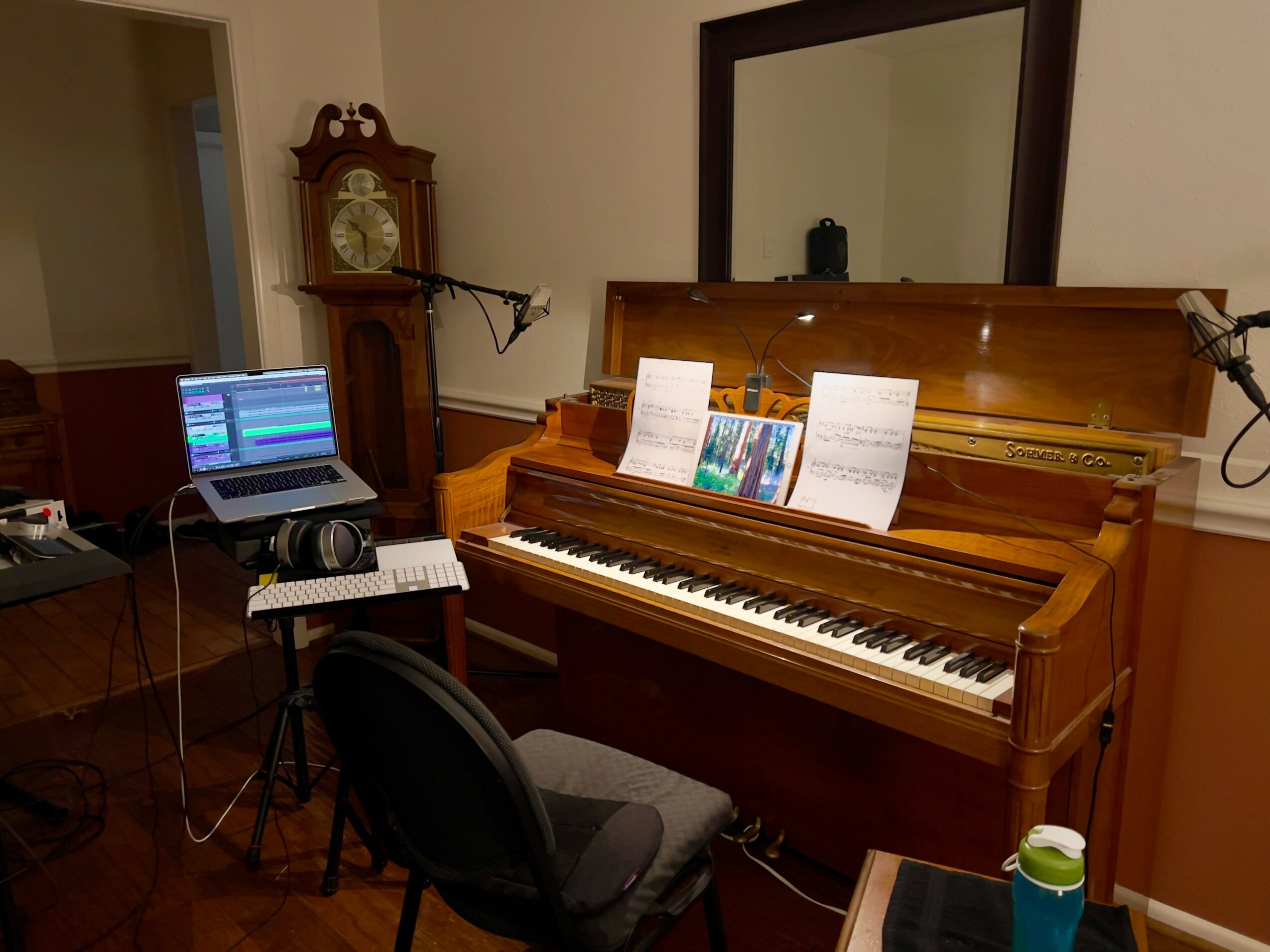Upright wooden piano with sheet music, a mirror above, and a lamp attached to the wall. A laptop and chair are nearby.