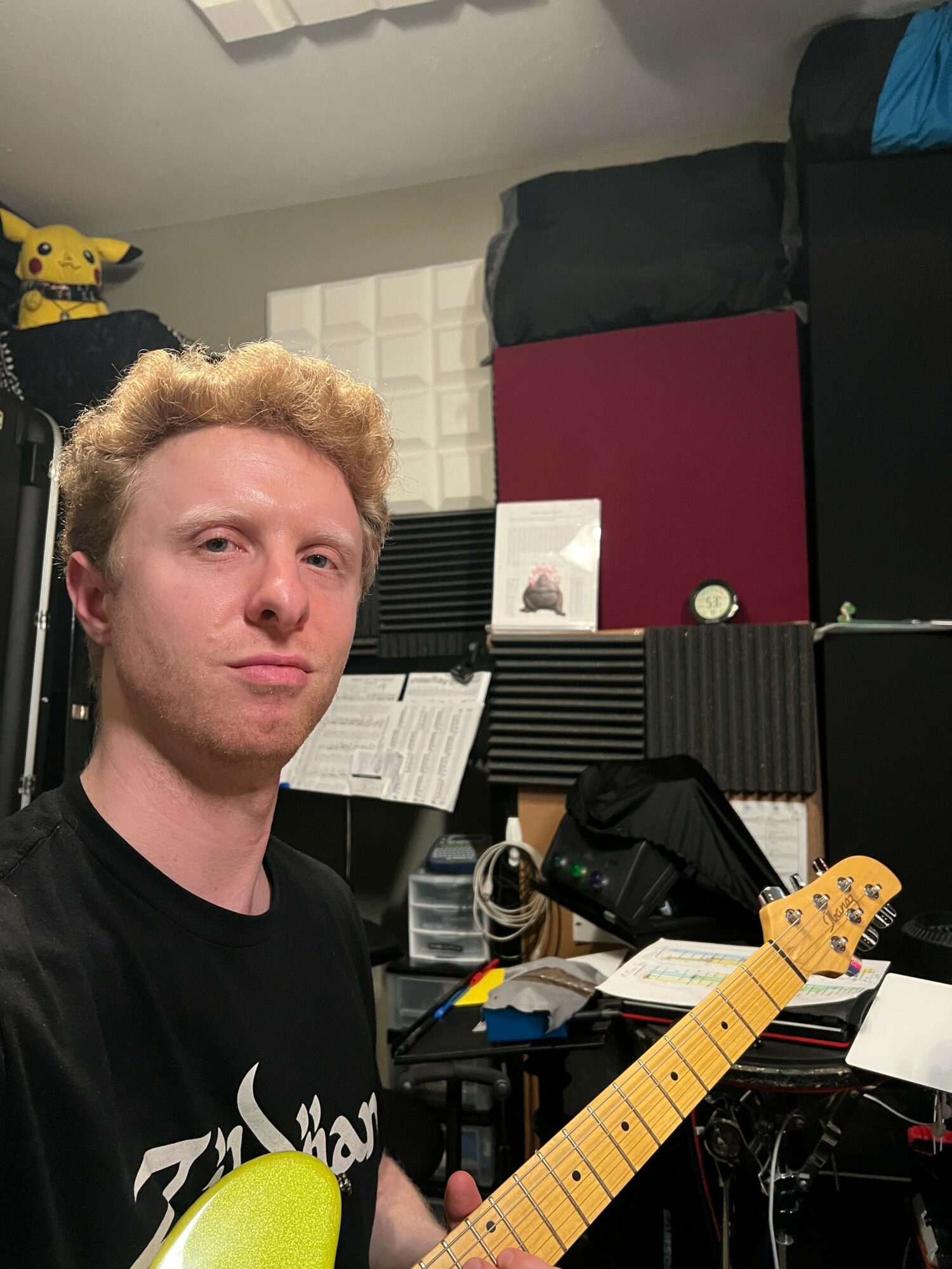 Young man with curly blonde hair in a music studio, holding a guitar neck, surrounded by music equipment and soundproofing panels.