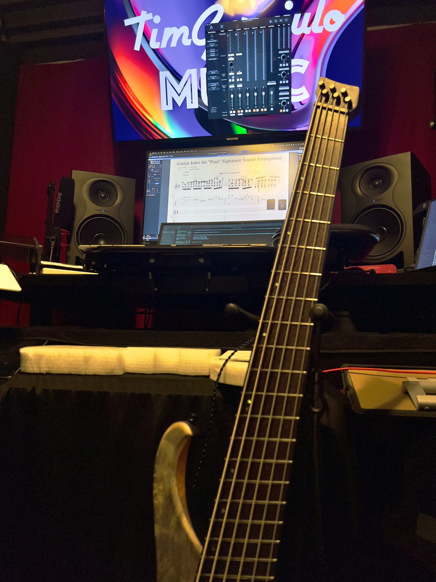 Electric guitar with a dark body and neck, in a music studio with computer screens and speakers.