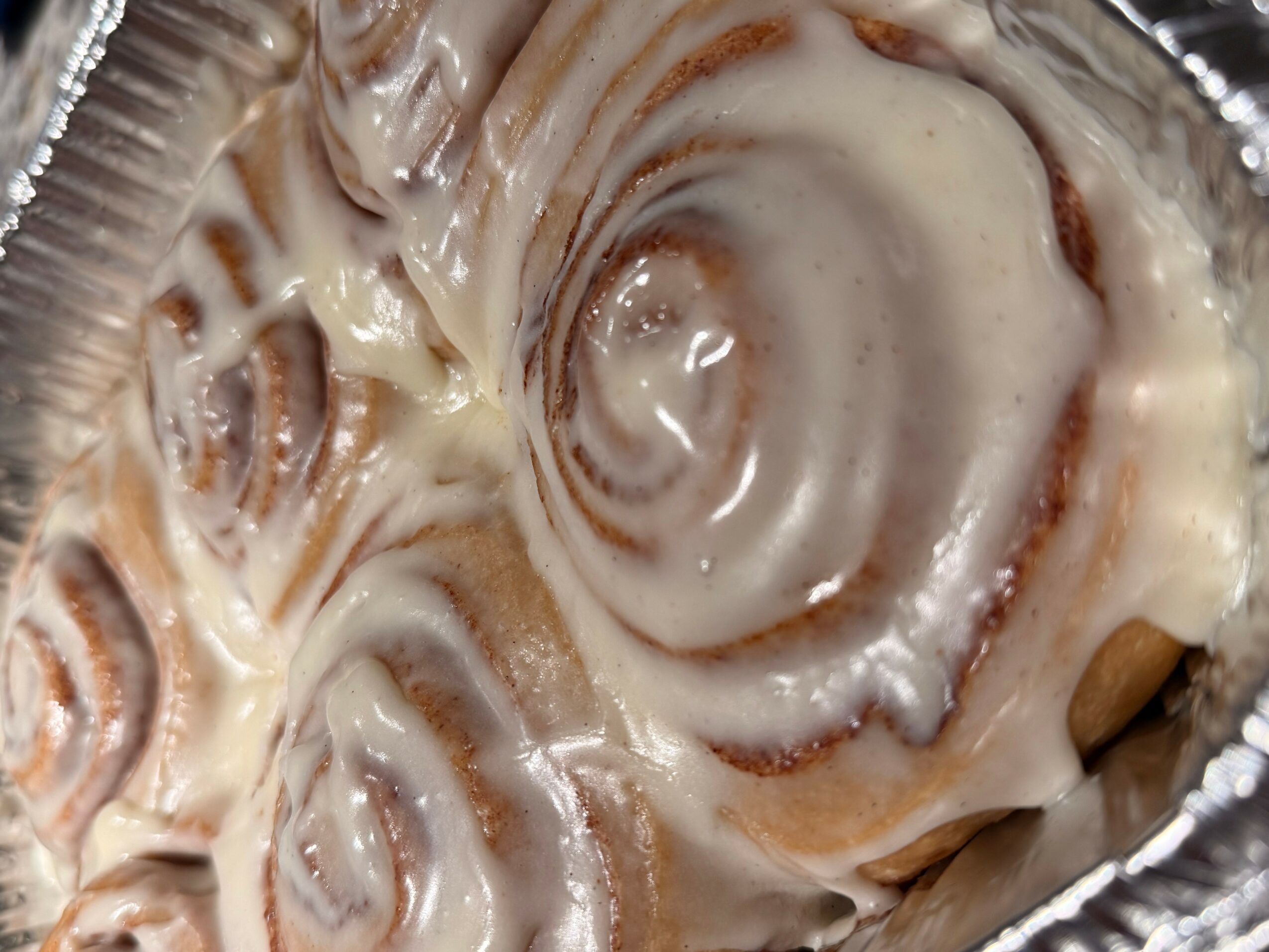 Close-up of a cinnamon roll with icing in a foil container, showing swirls and icing details.