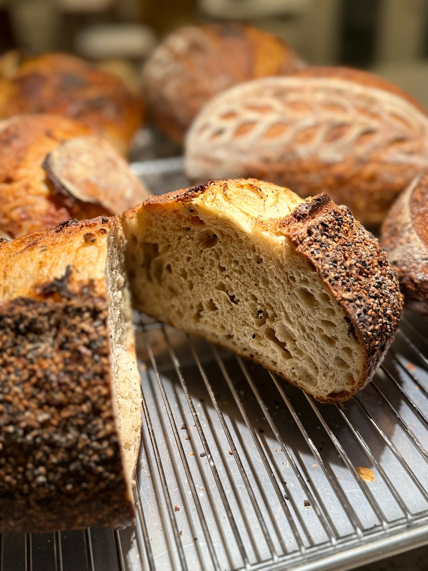 Sliced bread with a golden crust and airy interior, surrounded by various whole loaves on a cooling rack.