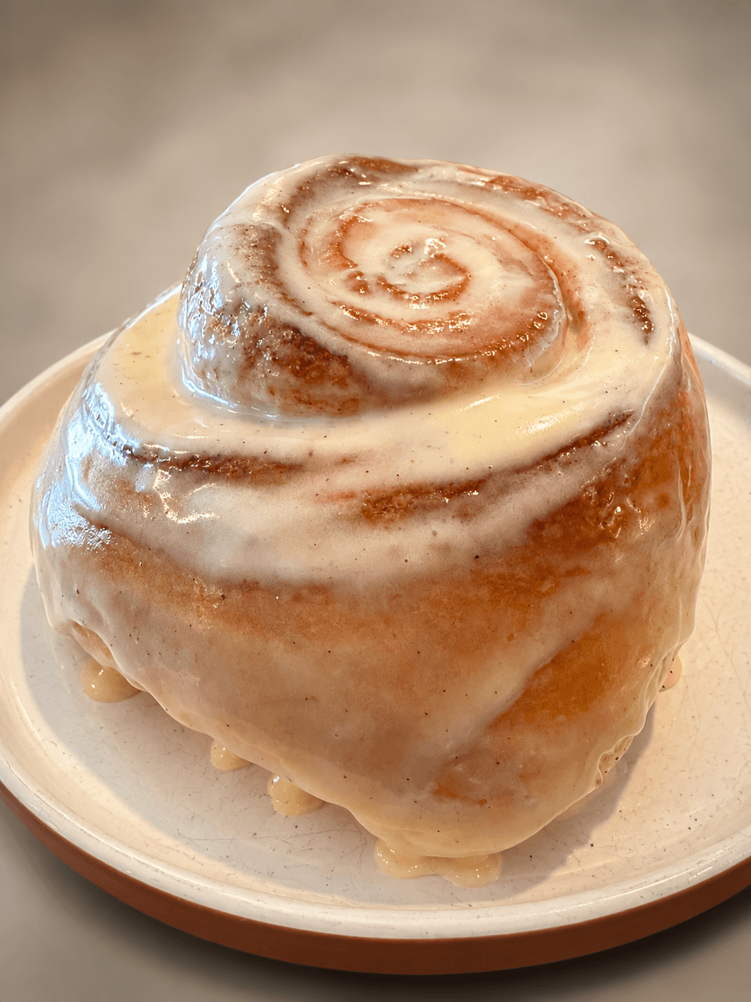 Cinnamon roll with icing on a small plate, viewed from the side, with a blurred background.