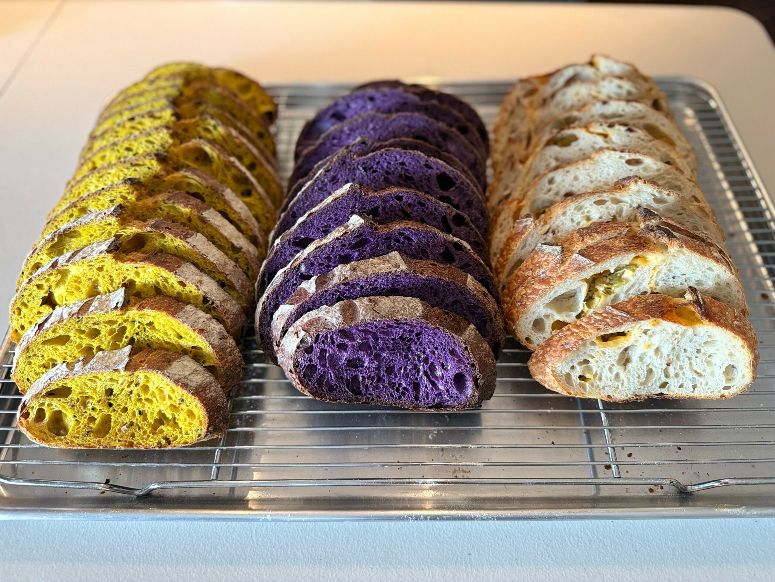 Three rows of sliced bread with different colors: yellow, purple, and light brown, on a cooling rack.