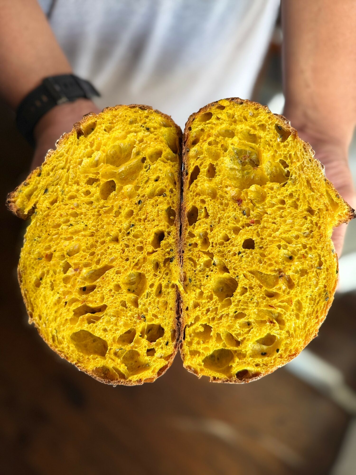 Person holding a large, yellow, airy bread with a textured interior, showing holes and a crust, in front of their torso.