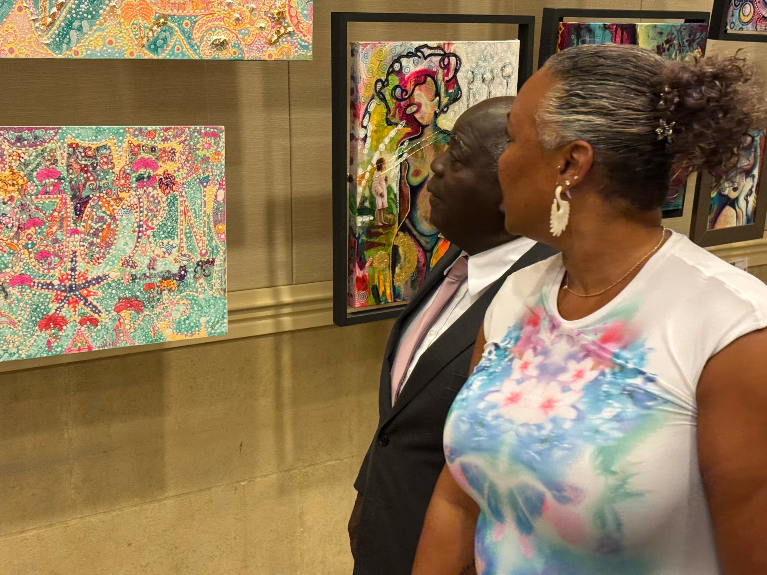 Two women look at colorful artwork in an art gallery, one wearing a white top and the other in a suit.