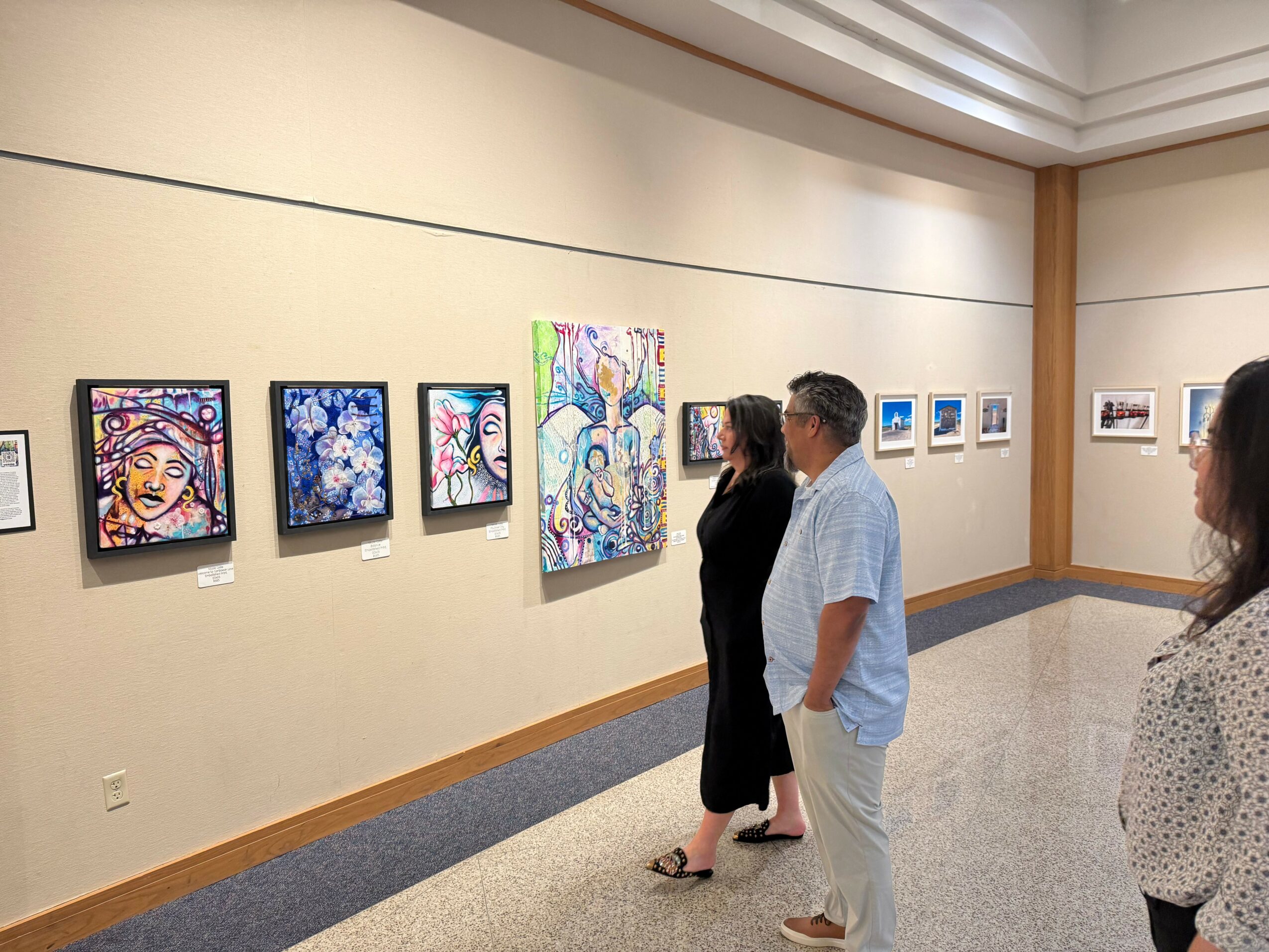 Three people observe colorful abstract paintings on a gallery wall, with additional artworks visible in the background.
