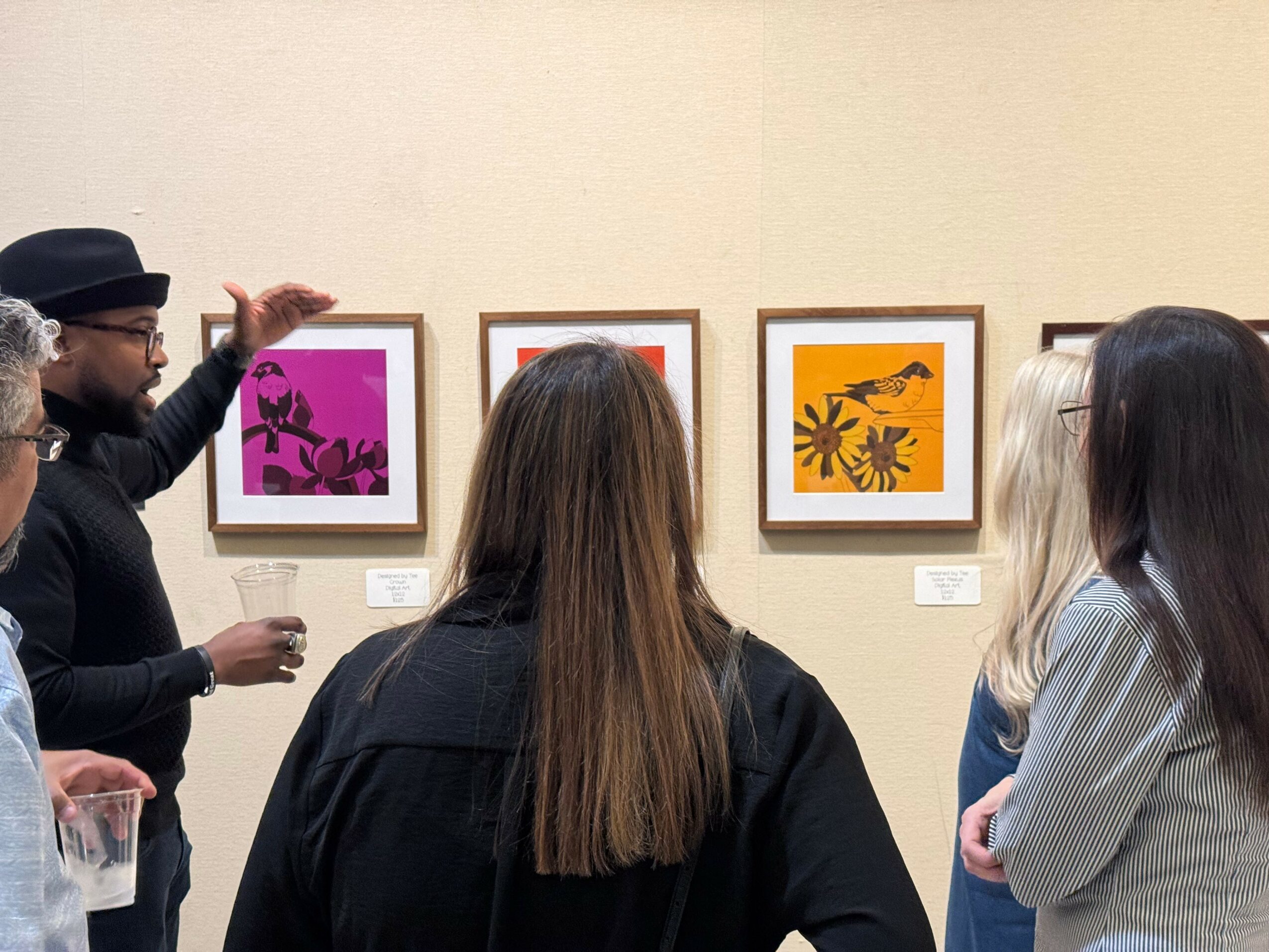 Group of people viewing framed artwork on a wall in an art gallery, with a person explaining the pieces.