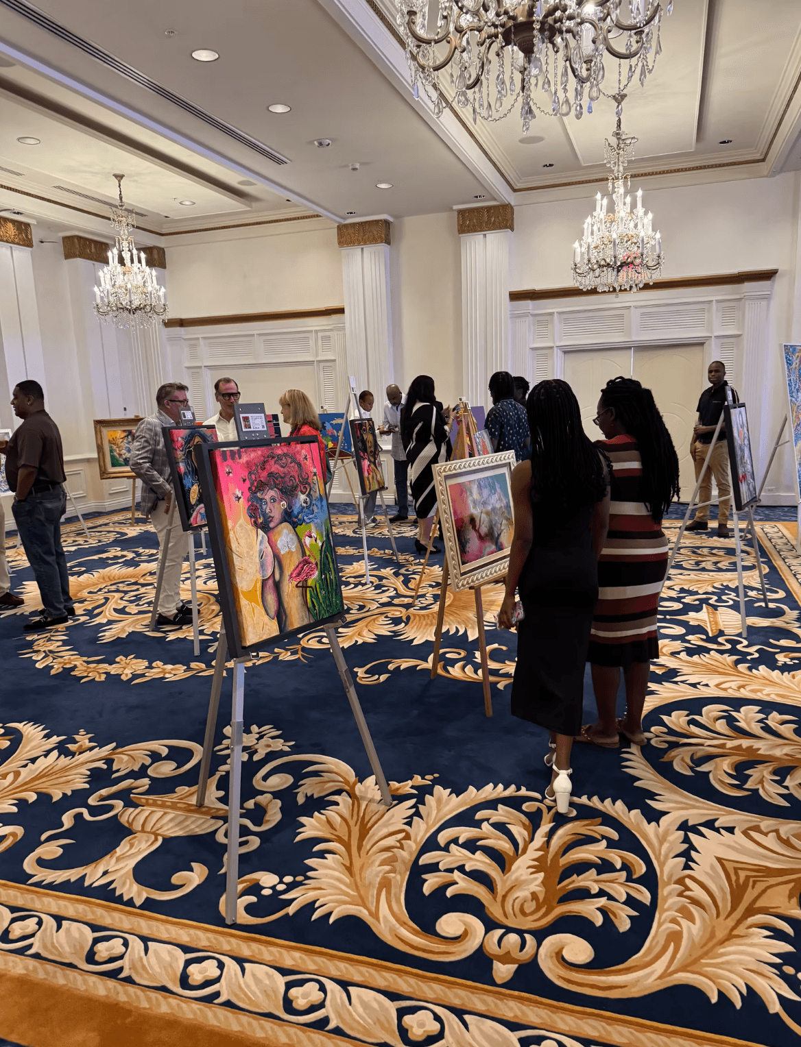 People viewing colorful paintings on easels in a spacious, ornate room with chandeliers and patterned carpet.