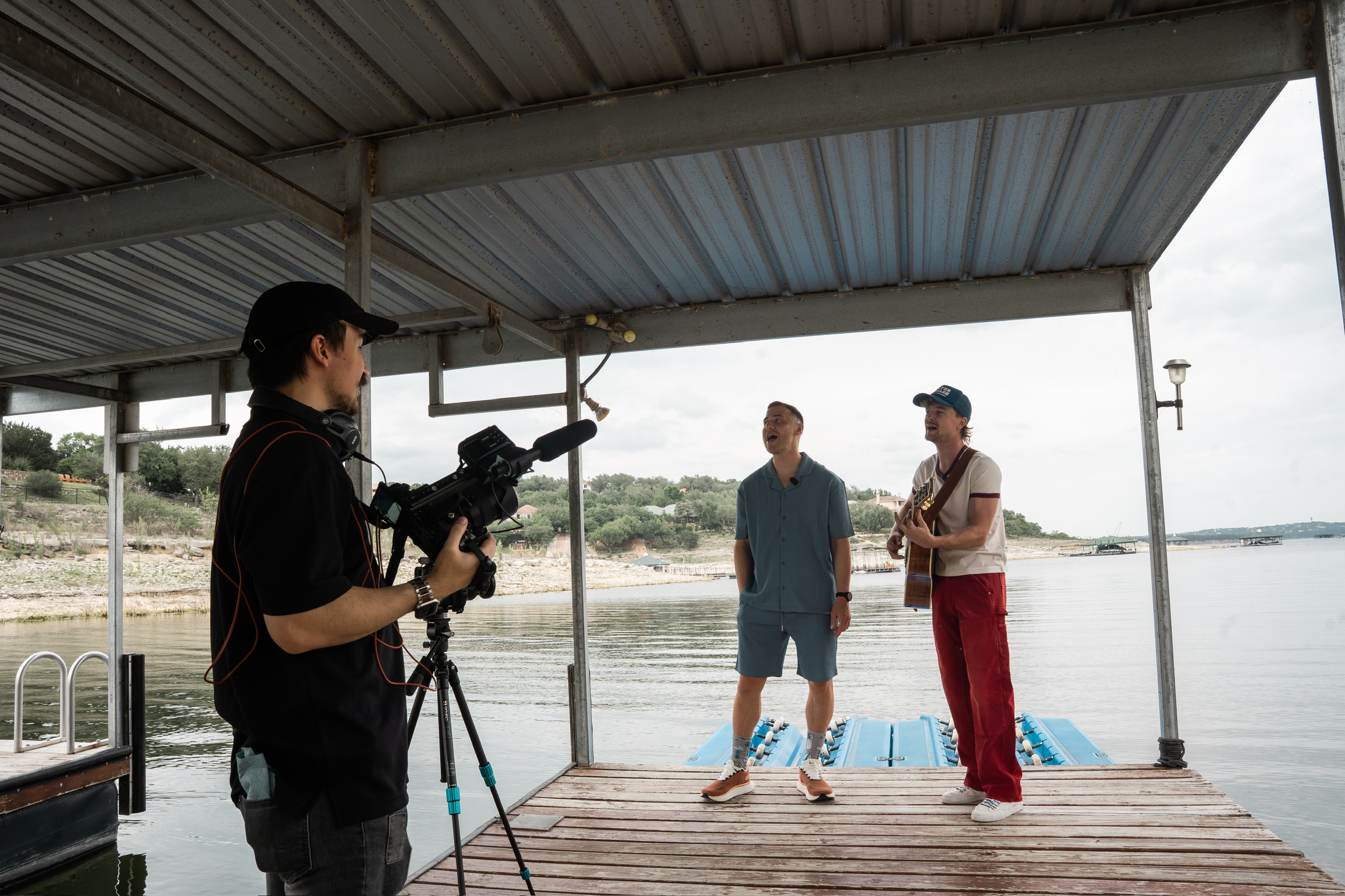 Two people stand on a dock by a body of water, with a person filming them under a metal roof structure.