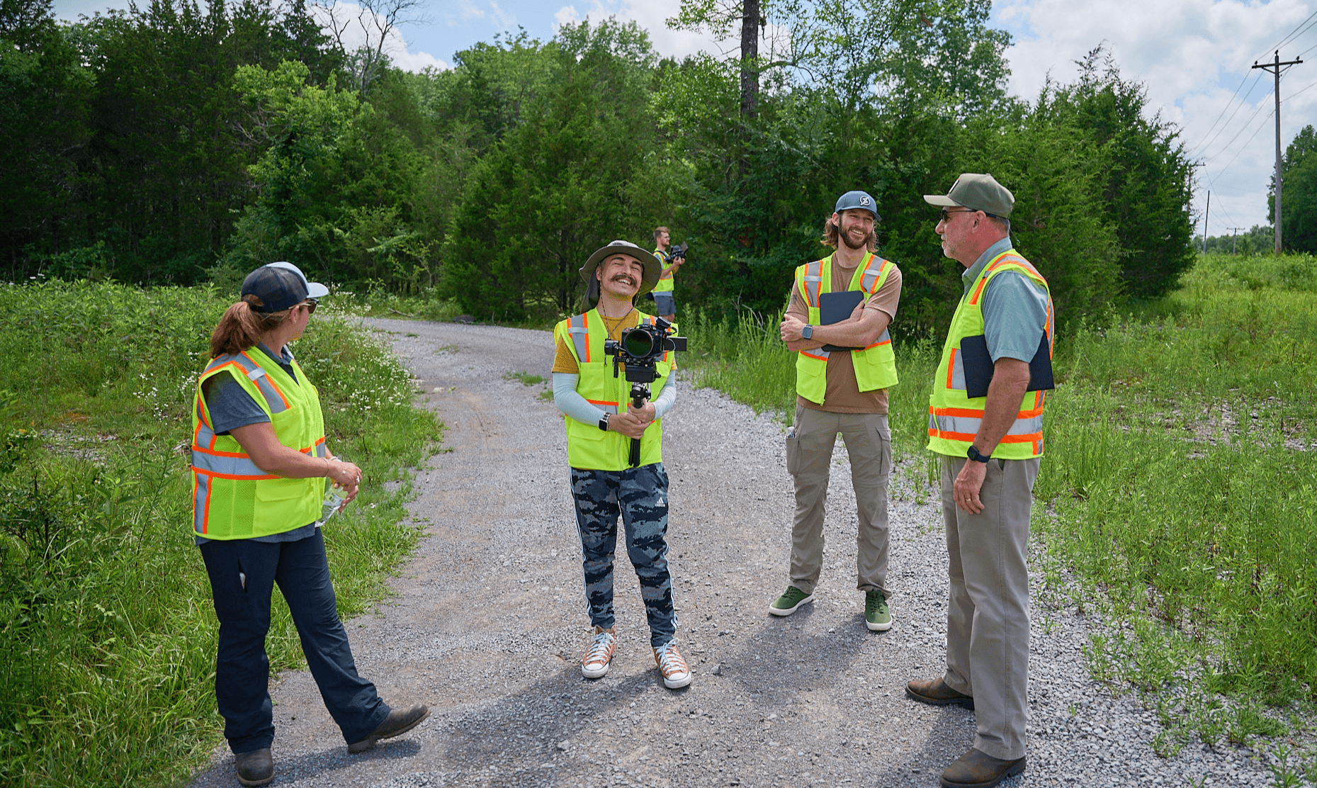 Four people wearing safety vests and hats standing on a gravel path outdoors, engaged in conversation, with greenery around.