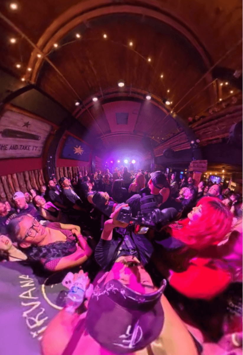 Crowd watching a stage with bright lights in a wooden venue, some people taking photos, colorful lighting, and a flag on the wall.