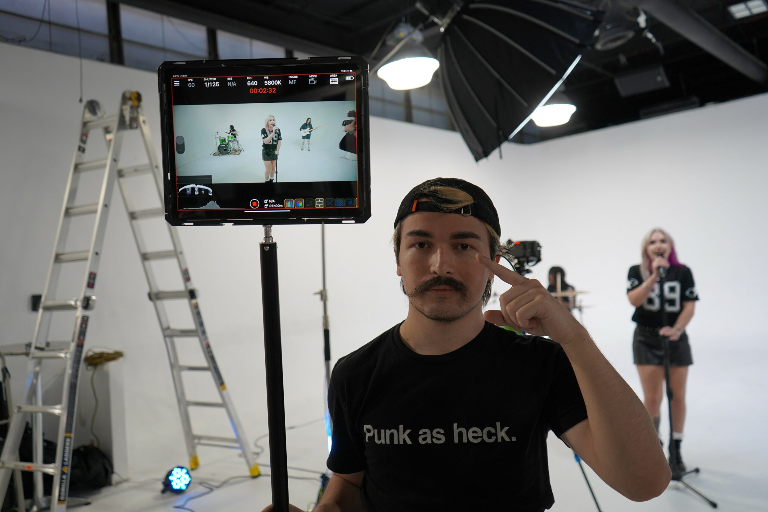 Man with a mustache and black T-shirt in a photography studio, holding a phone to his ear, with a woman in the background.