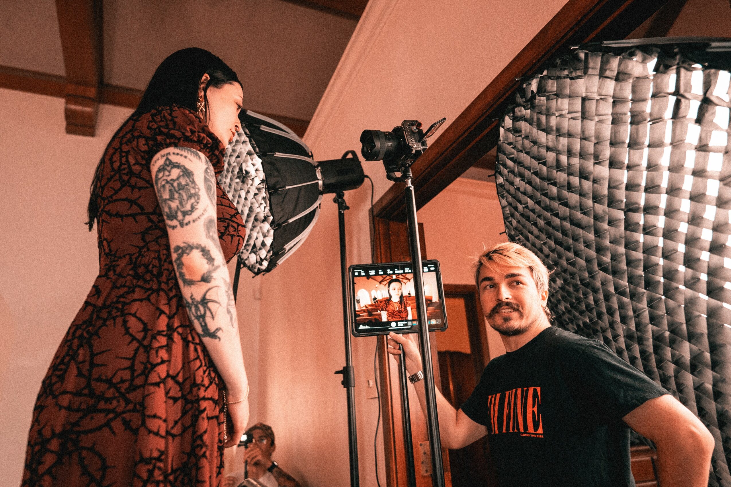A woman with tattoos on her arm stands next to a man with a beard, filming with a smartphone on a tripod inside a room.