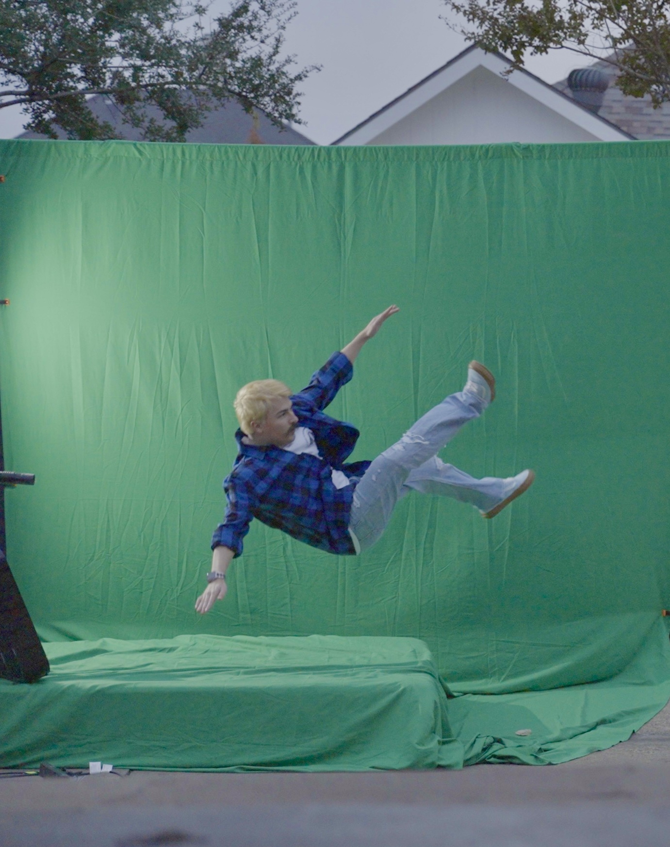 Child mid-air in front of a green screen, arms and legs extended, outdoors with trees and house in background.
