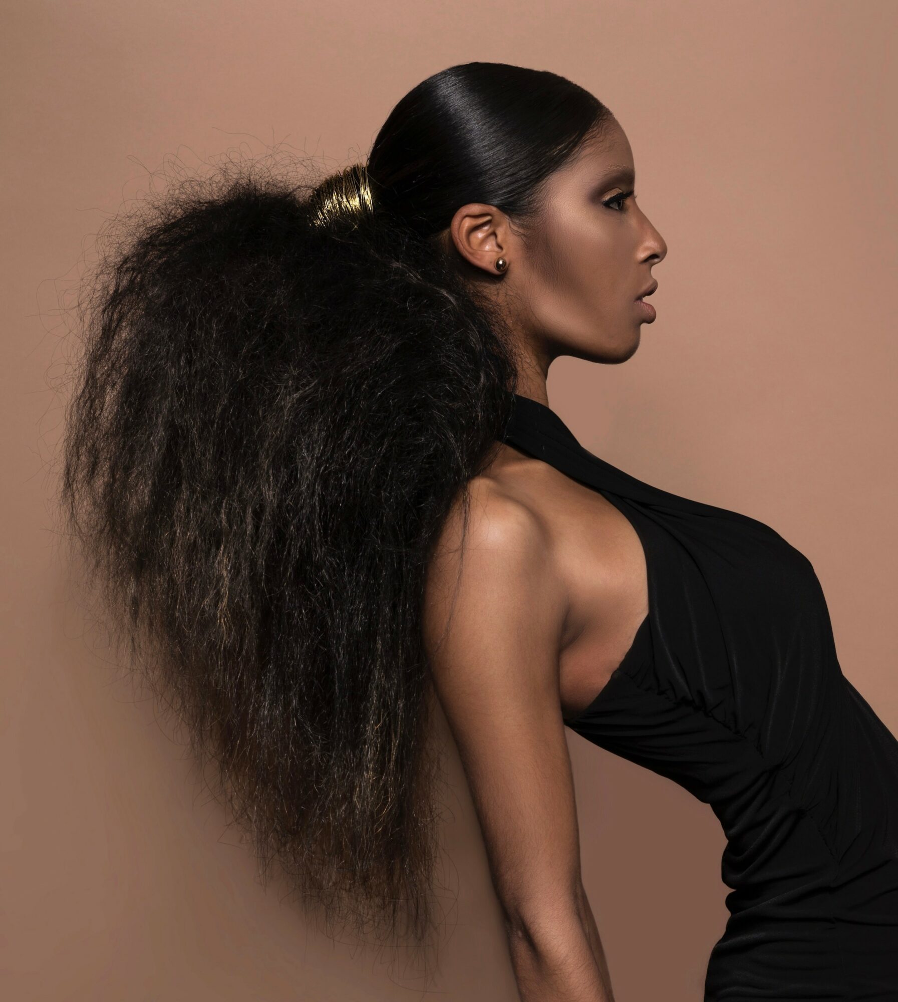 Side profile of a woman with long, voluminous curly hair tied back, wearing a black sleeveless top, against a beige background.