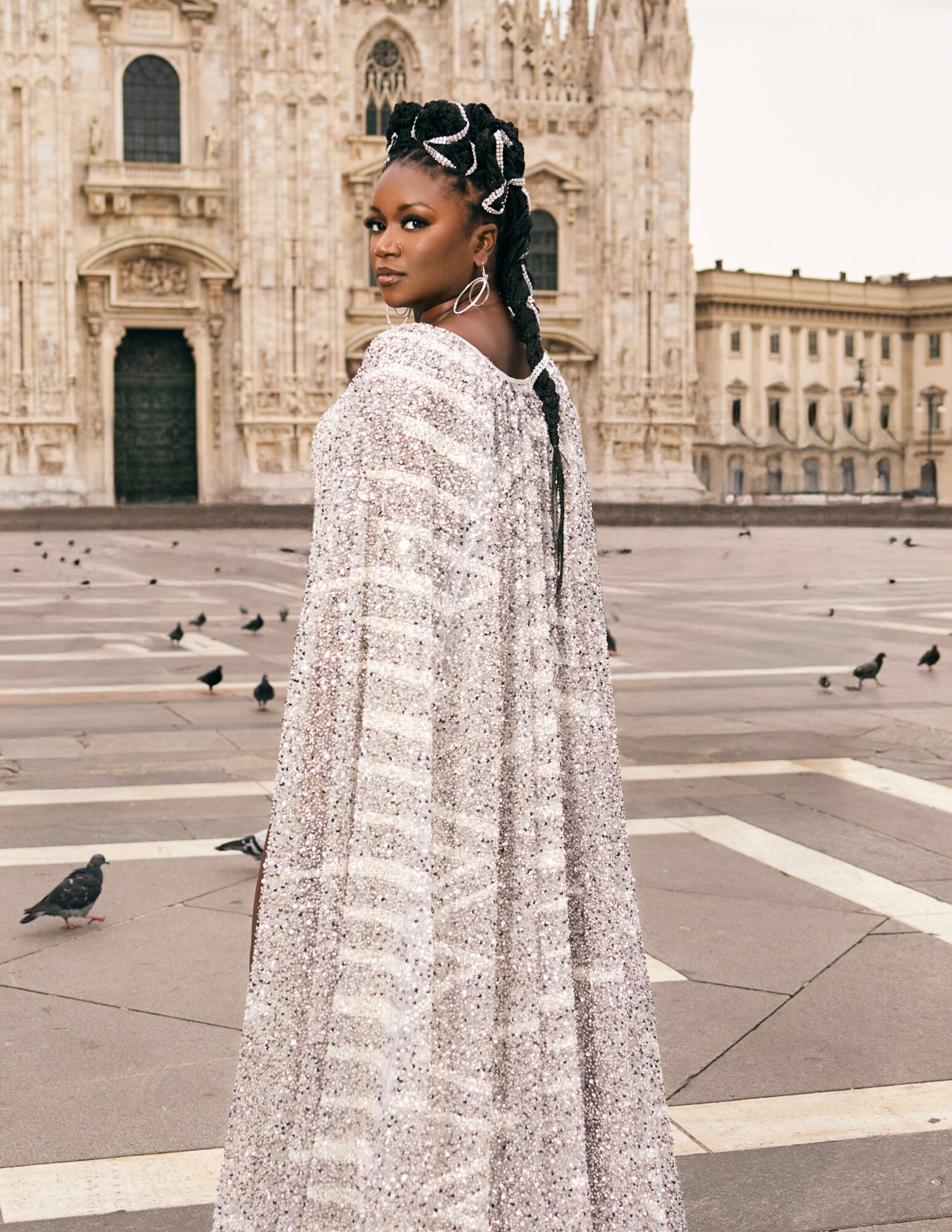 Woman with braided hair and headwrap standing in front of historic building, wearing a long, patterned dress, pigeons on ground.