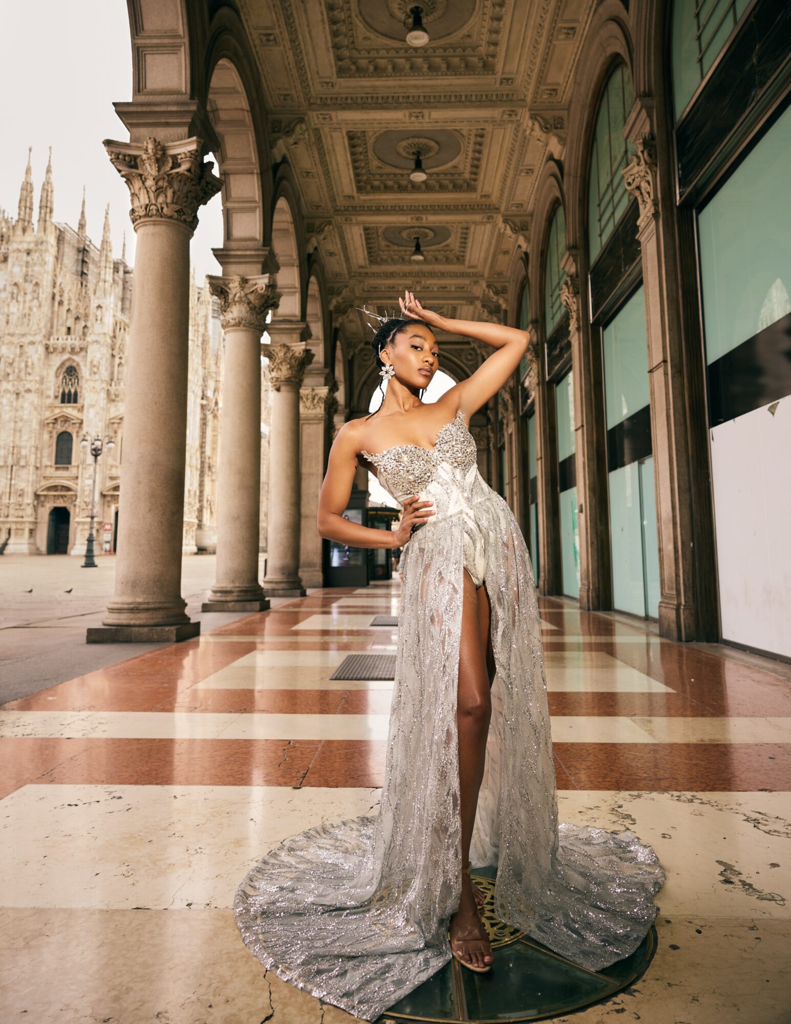 Woman in a silver gown with a slit, standing in an ornate corridor with arches and columns, looking into the distance.