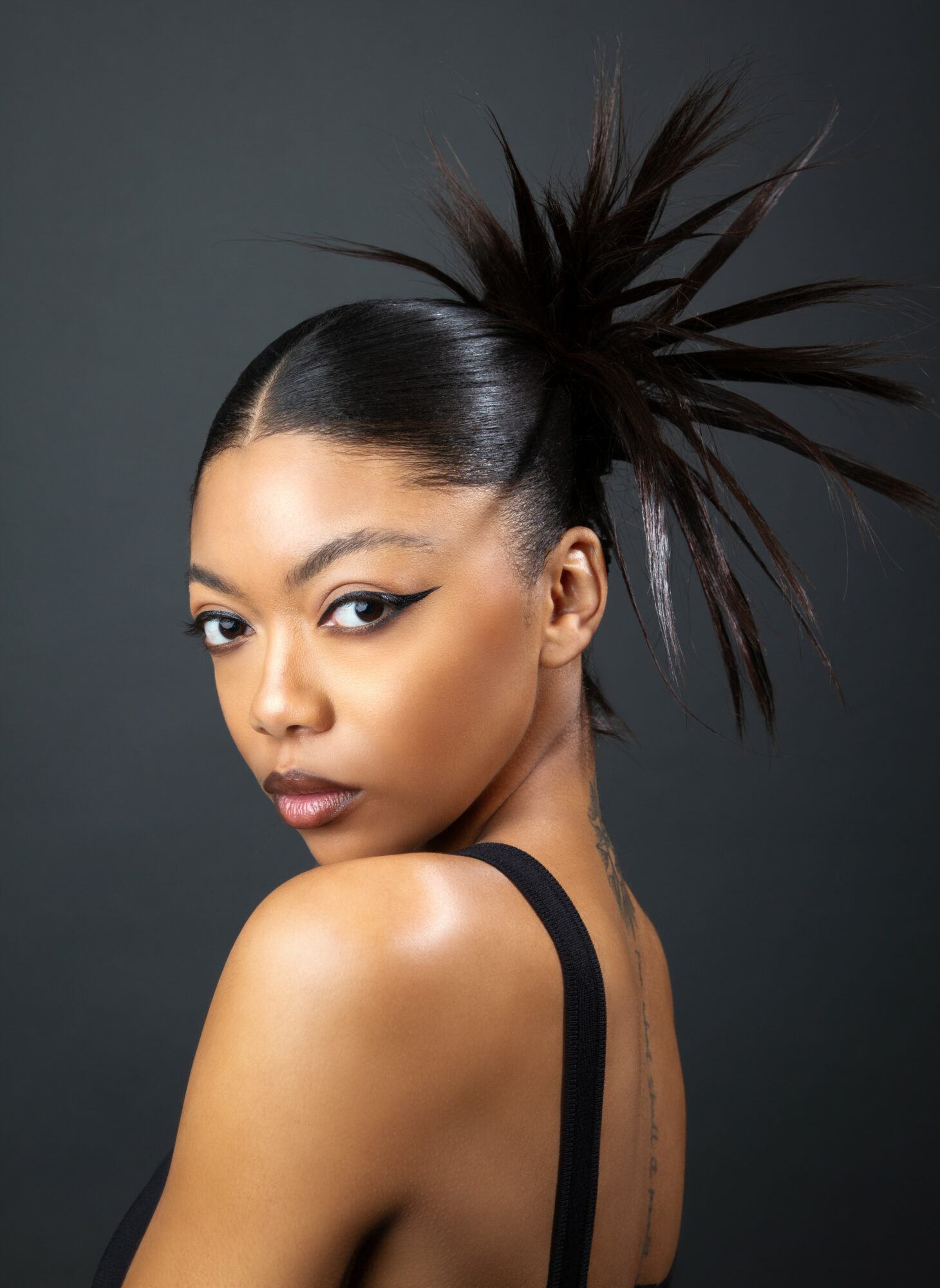 Woman with styled hair and makeup, looking over shoulder against dark background.