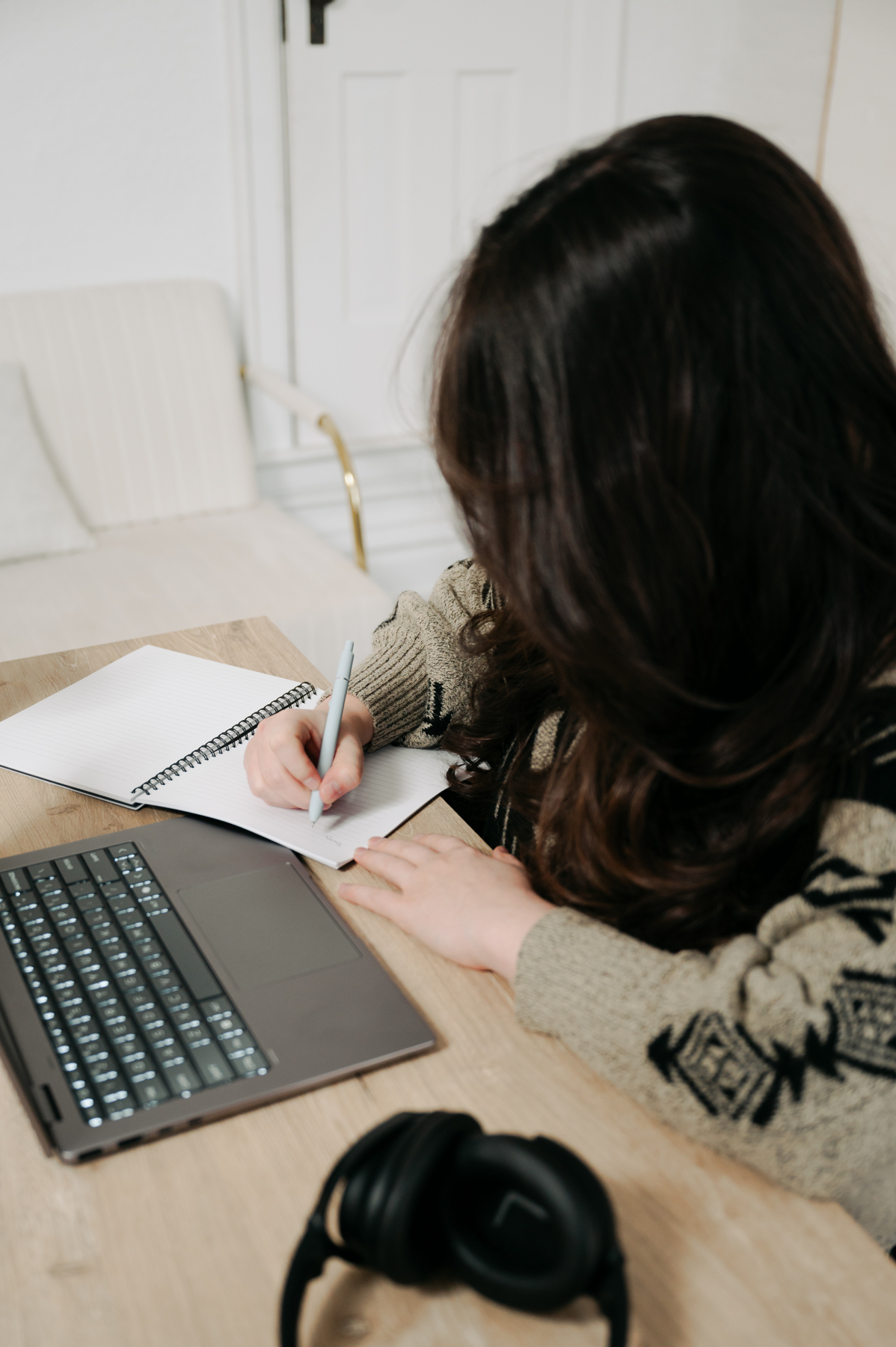 Person with dark hair writing in a notebook at a table with a laptop and headphones, in a bright room.