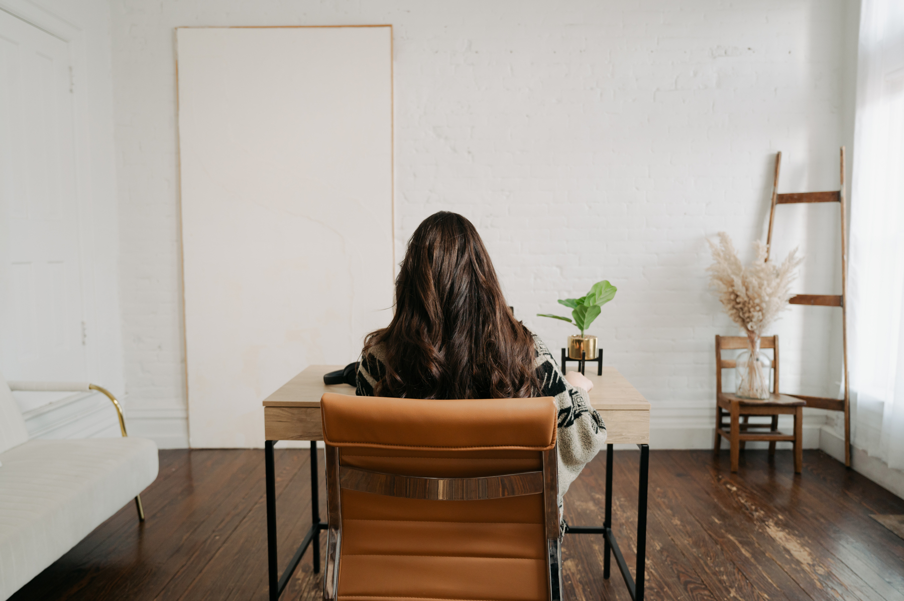 Person with long dark hair sitting at a desk in a bright room, facing away from the camera, with a chair and decor.