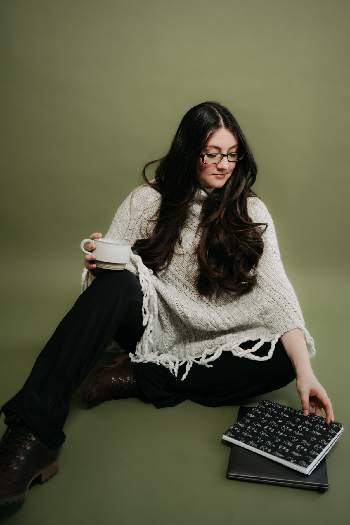 Woman with long dark hair and glasses sitting on the floor, holding a mug, with a keyboard and notebook nearby, against a green background.