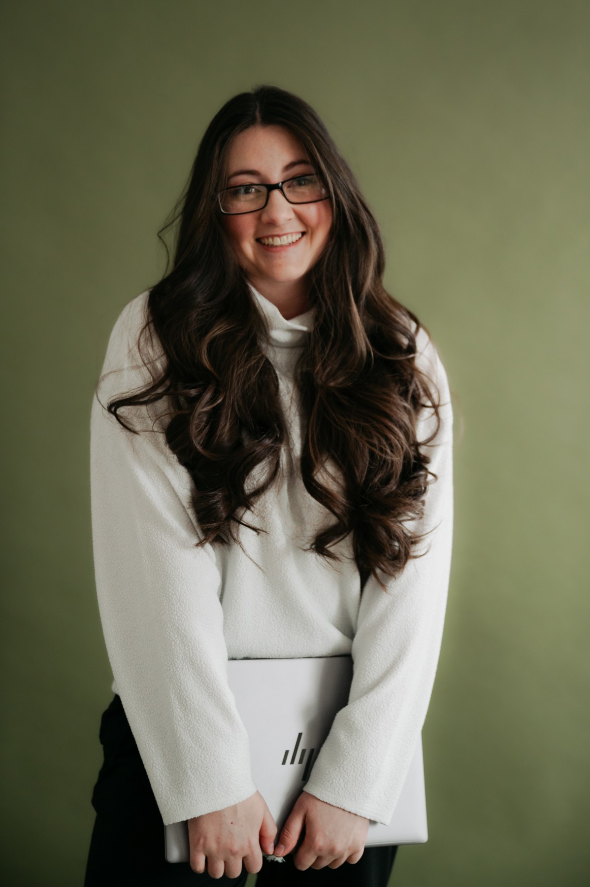 Young woman with long dark hair and glasses smiling, holding a closed laptop, standing against a green background.
