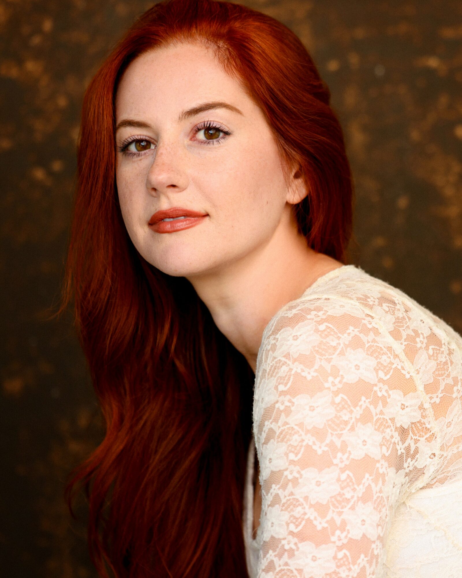 Portrait of a woman with long red hair wearing a white lace top, looking at the camera.