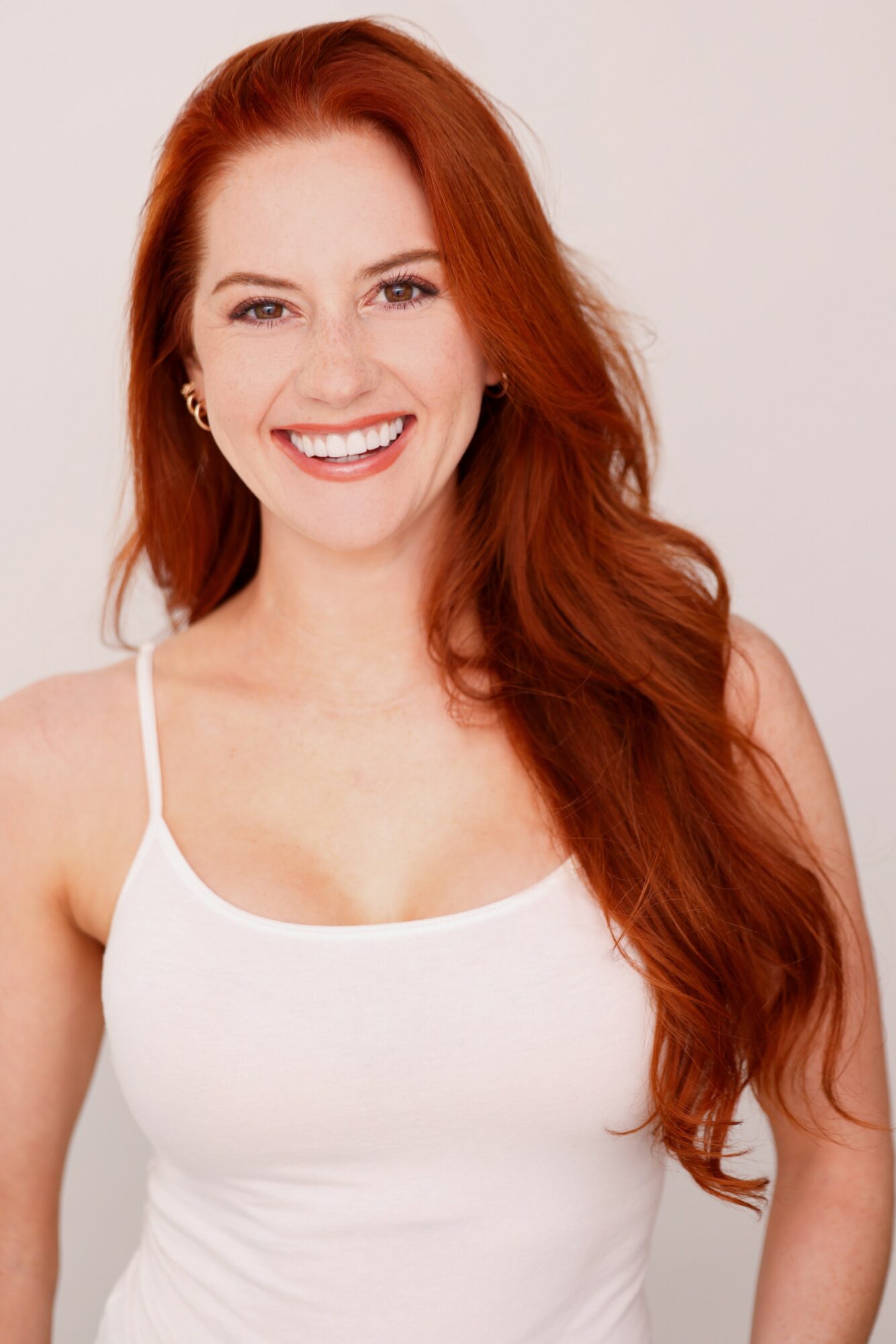 Smiling woman with long red hair wearing a white tank top, facing forward, against a plain background.