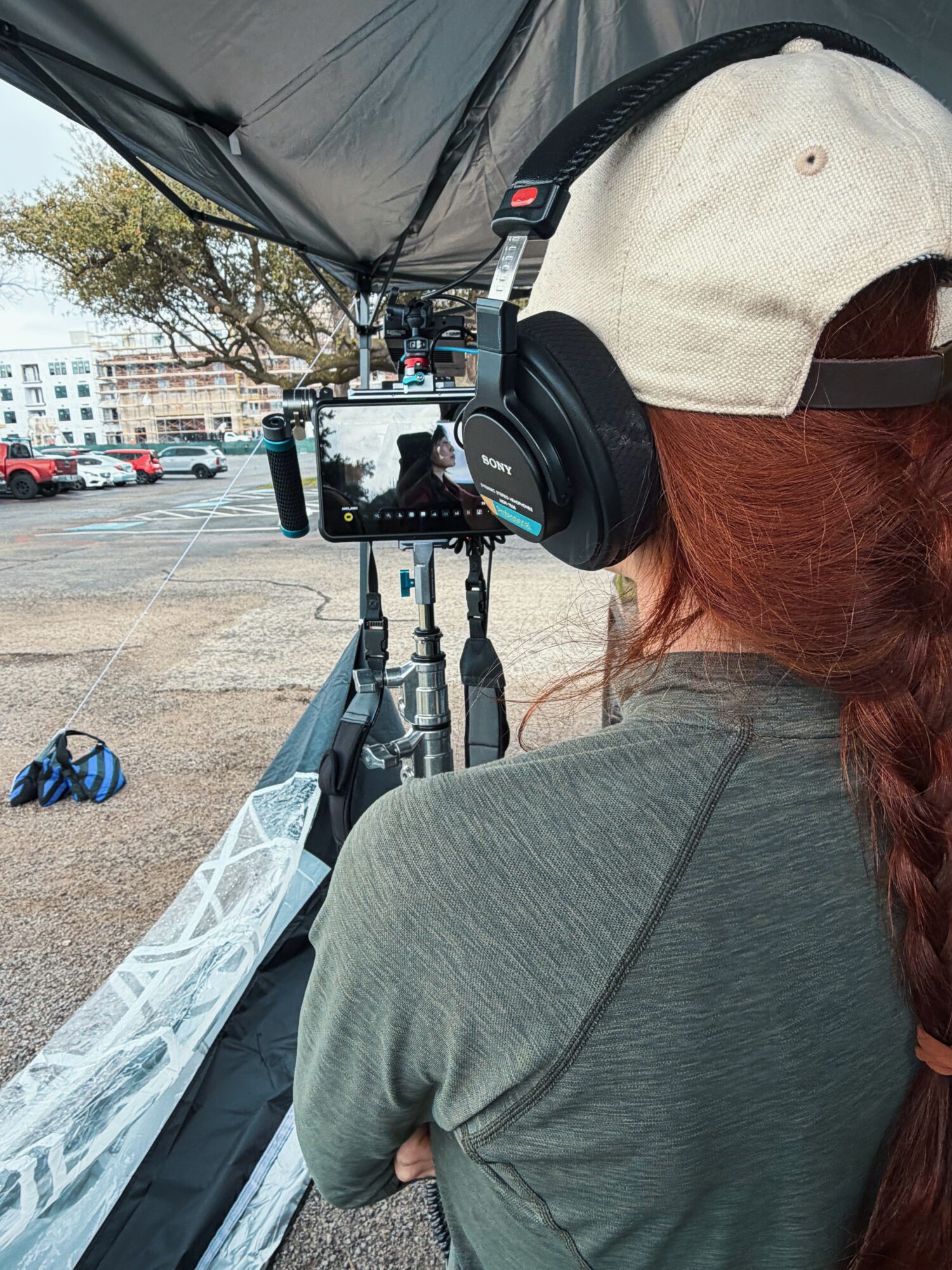 Person with long red hair, wearing a beige cap and headphones, operating camera equipment outdoors.