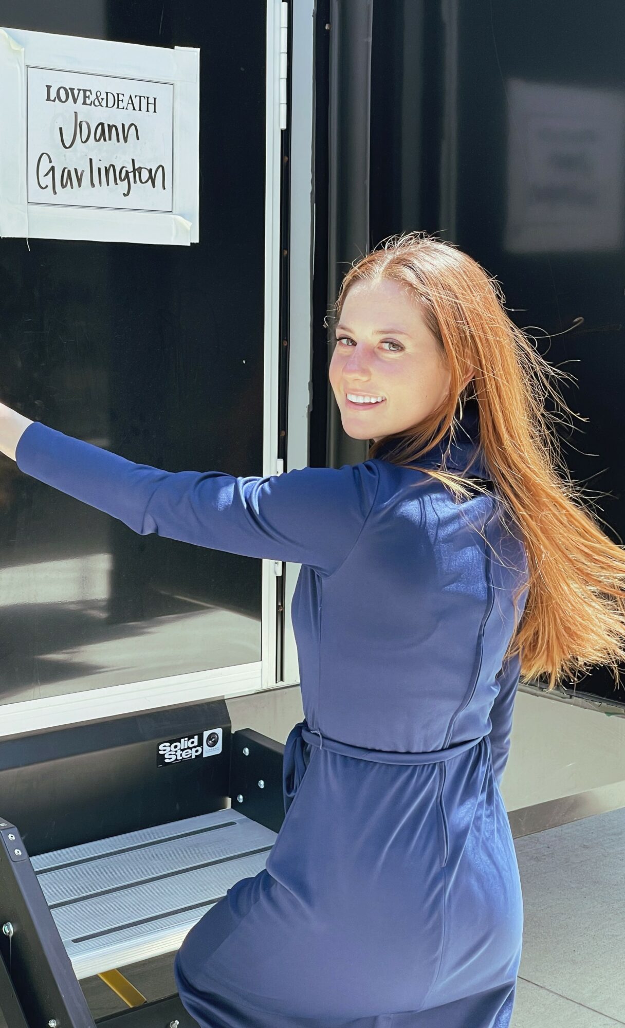Young woman with long red hair smiling and reaching out, wearing a blue dress, standing outside a glass door with a sign.