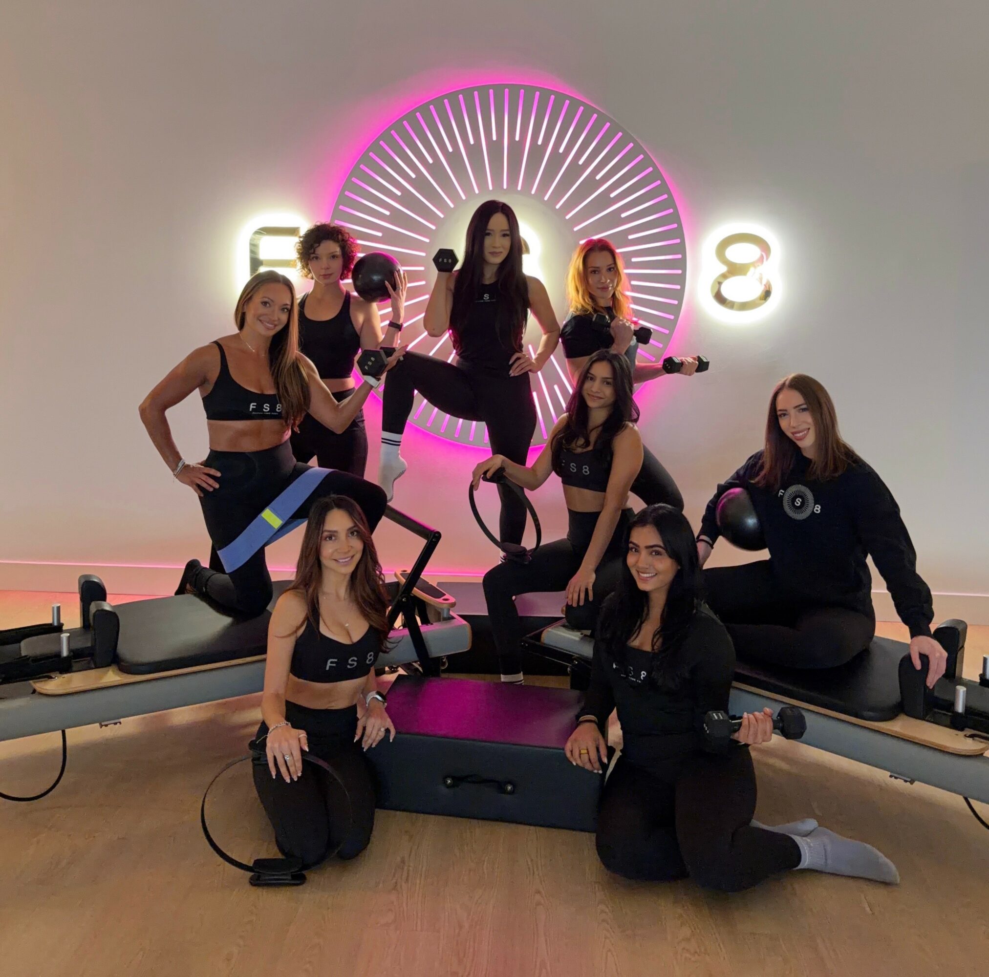 Group of women in workout attire posing with fitness equipment in a studio with pink and white neon lights, and a circular design on the wall.