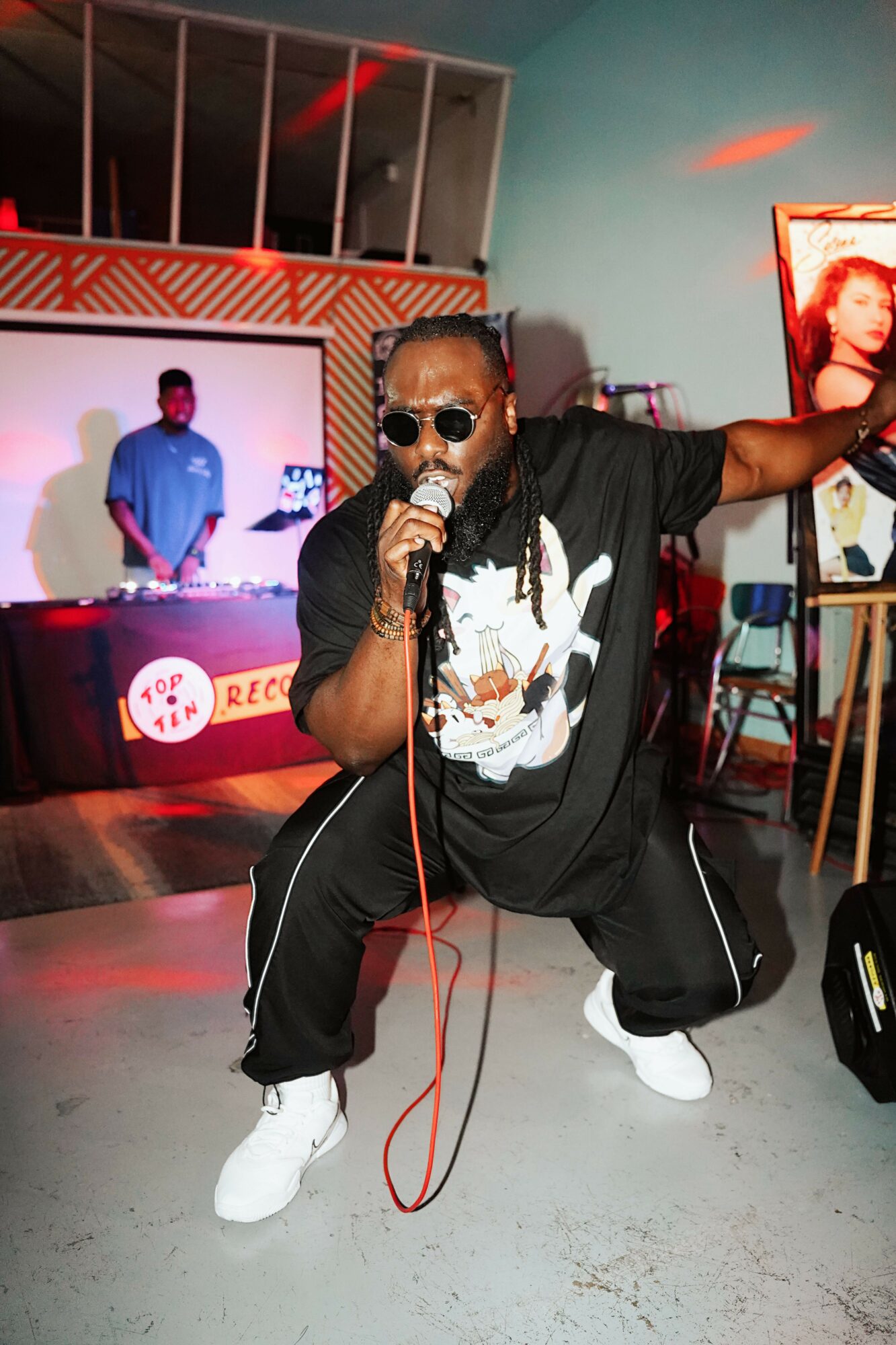 Person crouching with sunglasses, headphones, and a microphone in a club or event space, with a stage and screen in background.