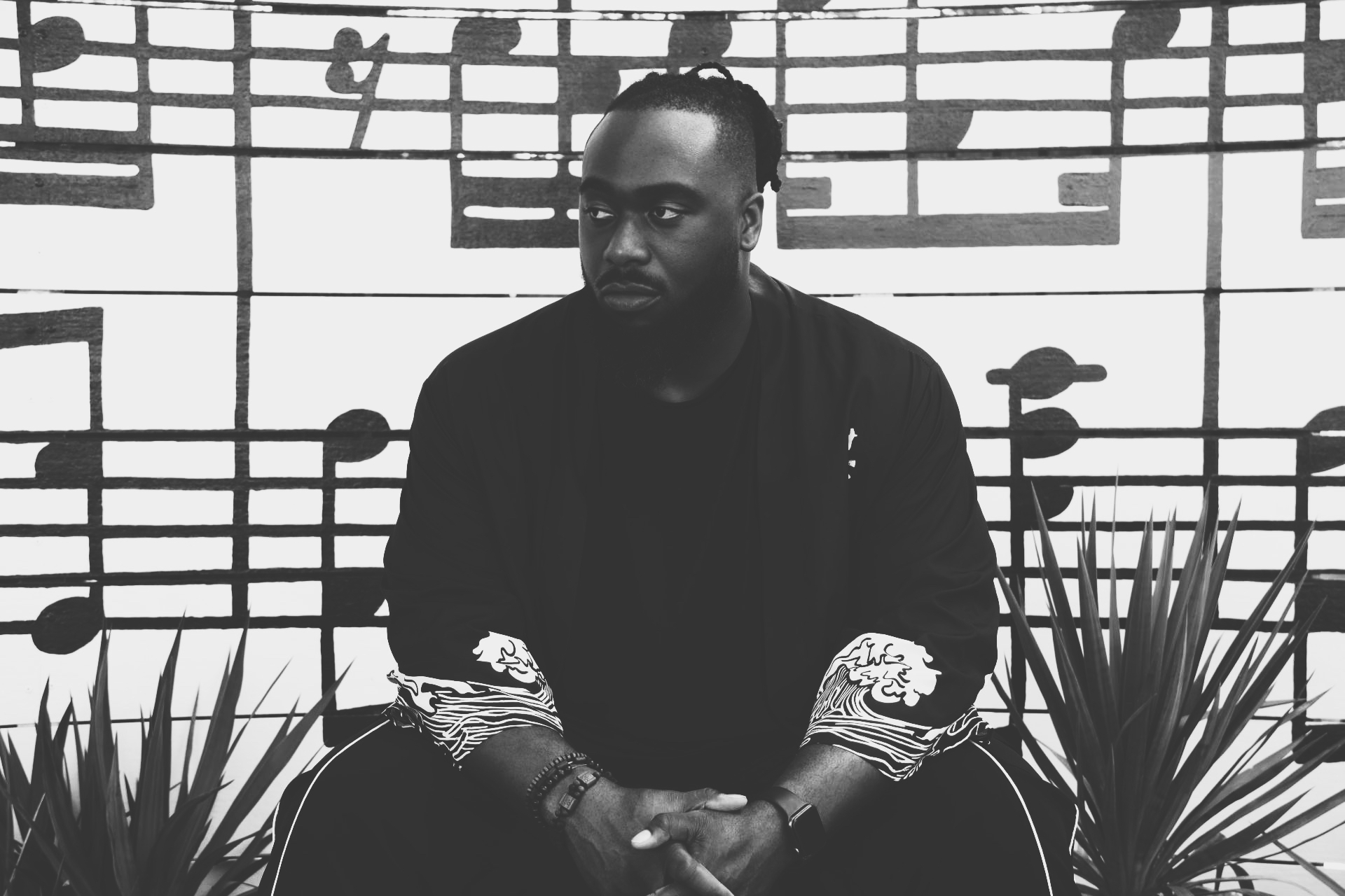 Black man sitting with hands clasped, background of musical notes on a wall, plants on sides, in black and white.