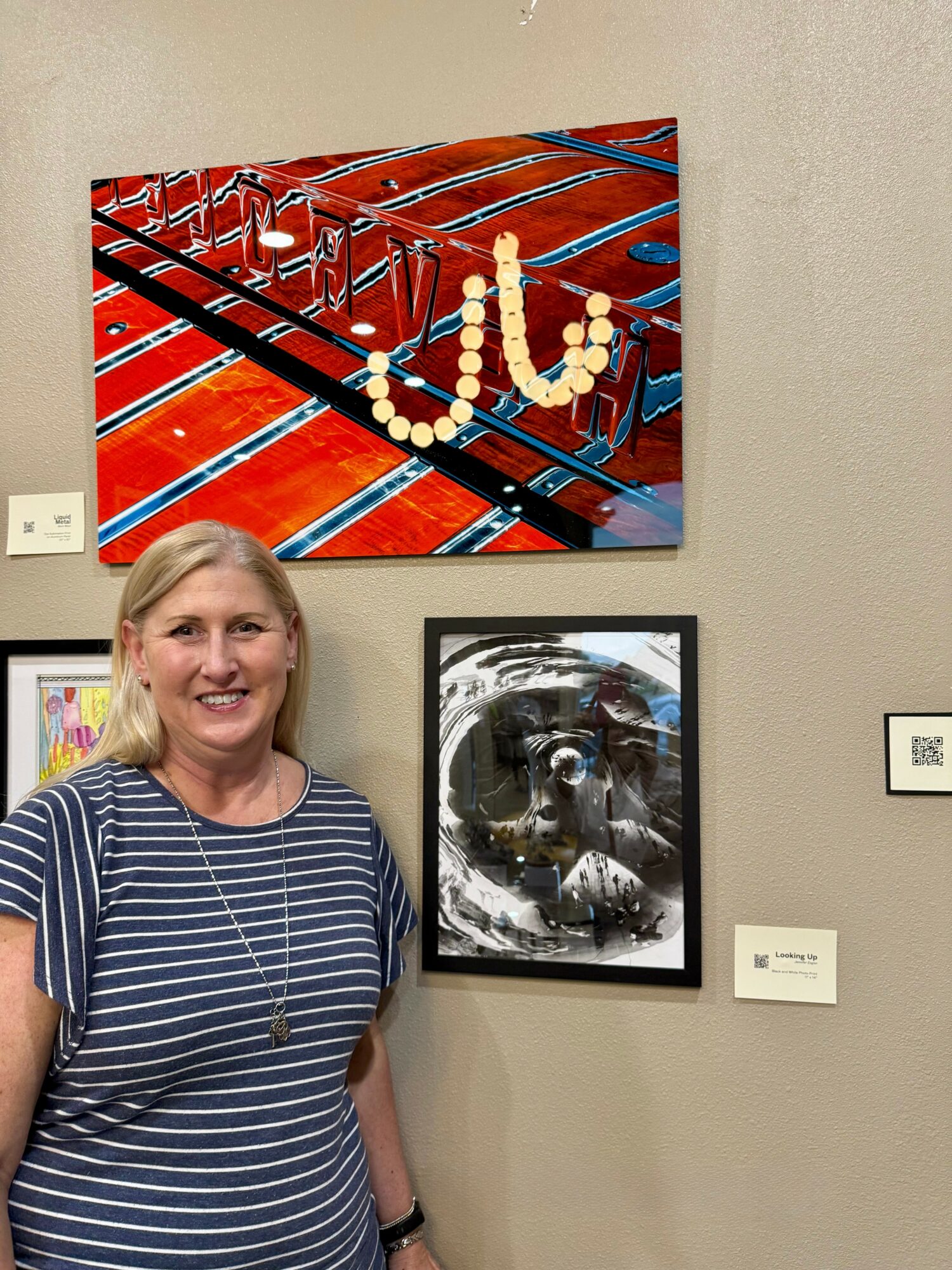 Smiling woman with blonde hair in a striped shirt standing in front of two framed artworks on a beige wall.