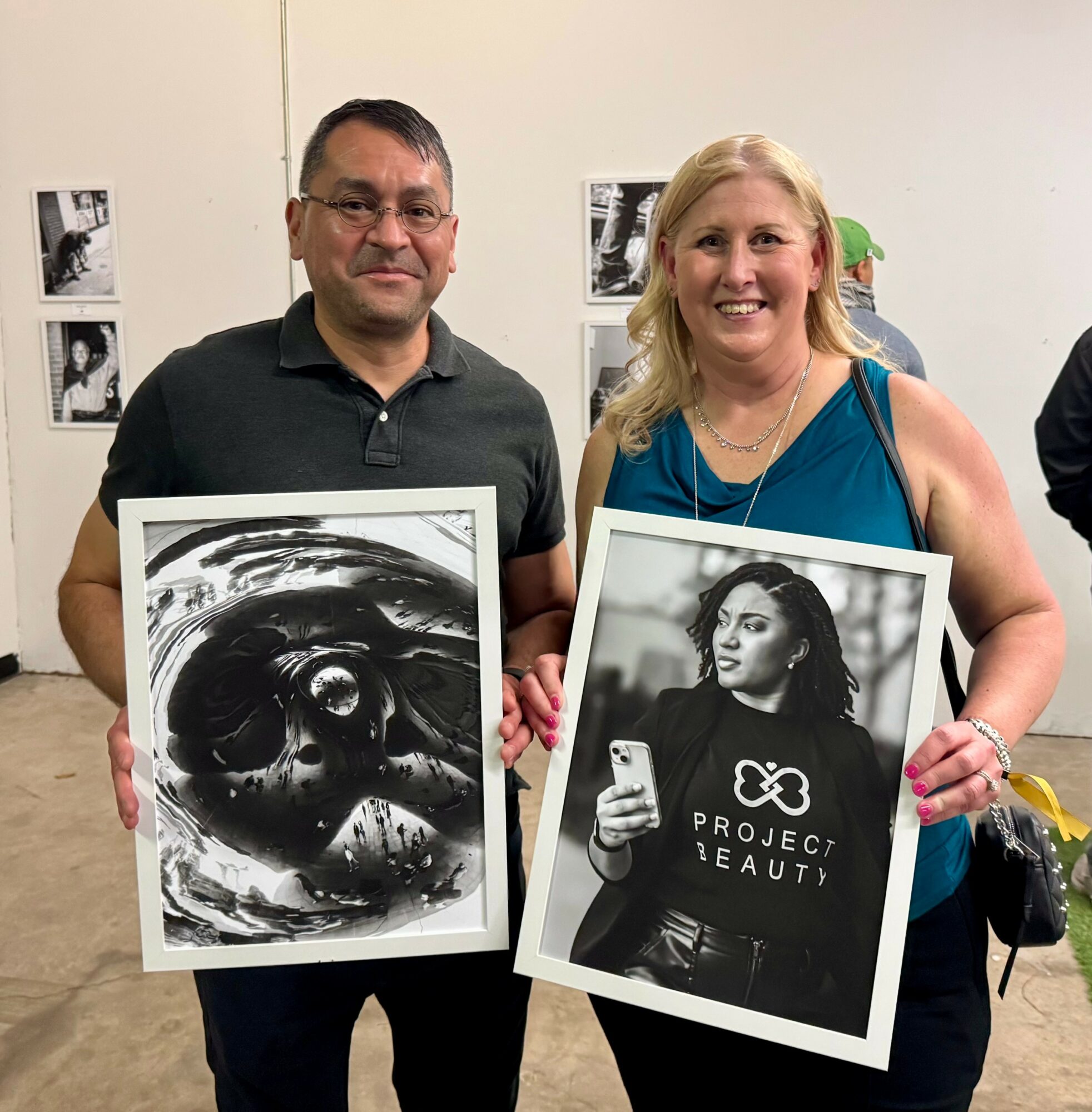 Two people holding framed black-and-white photographs, smiling, standing indoors with photos on the wall behind them.